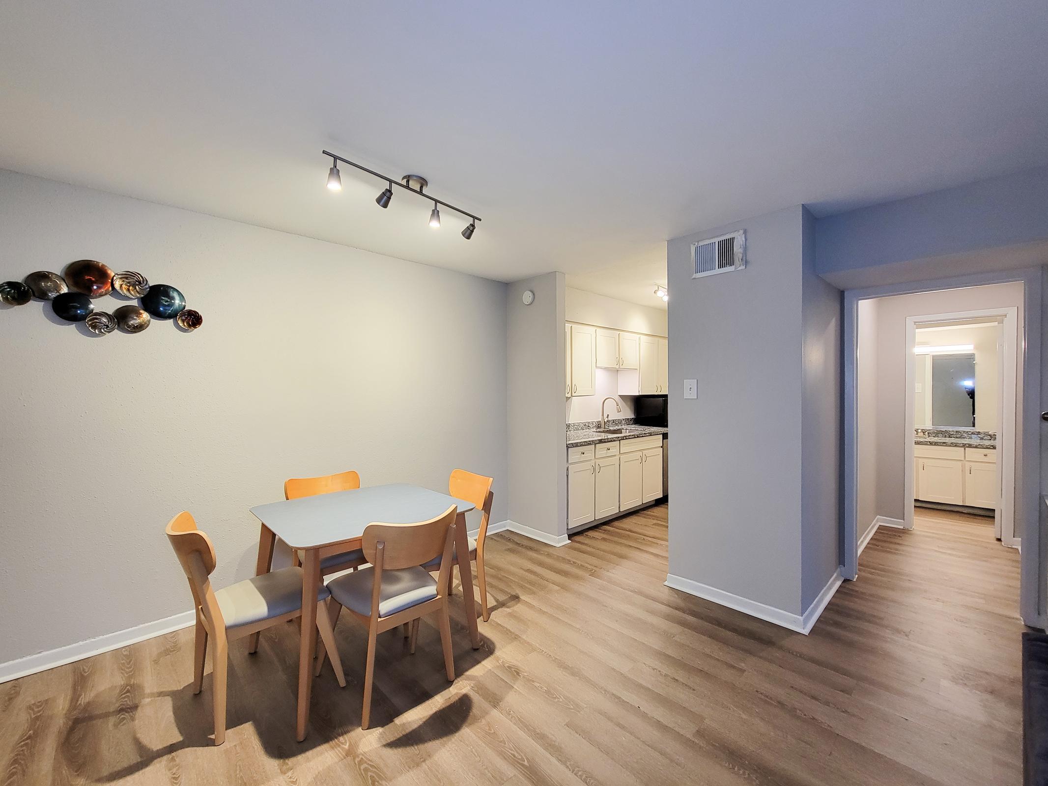 A modern dining area featuring a light-colored table with four wooden chairs, adjacent to a kitchen with white cabinetry. The walls are painted light gray, and there is a decorative wall piece with circular plates. A hallway leading to another room is visible in the background, with natural light coming from a window.
