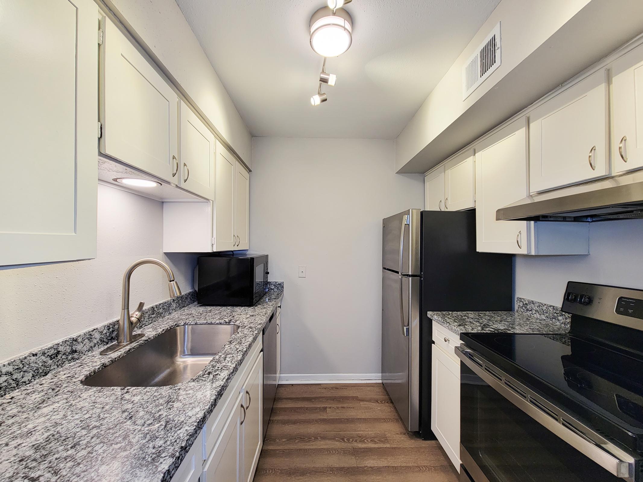 A modern kitchen featuring white cabinets, a granite countertop, a stainless steel sink with a gooseneck faucet, a black microwave, and an electric stove. The space includes a stainless steel refrigerator, under-cabinet lighting, and wooden flooring, creating a bright and functional cooking area.