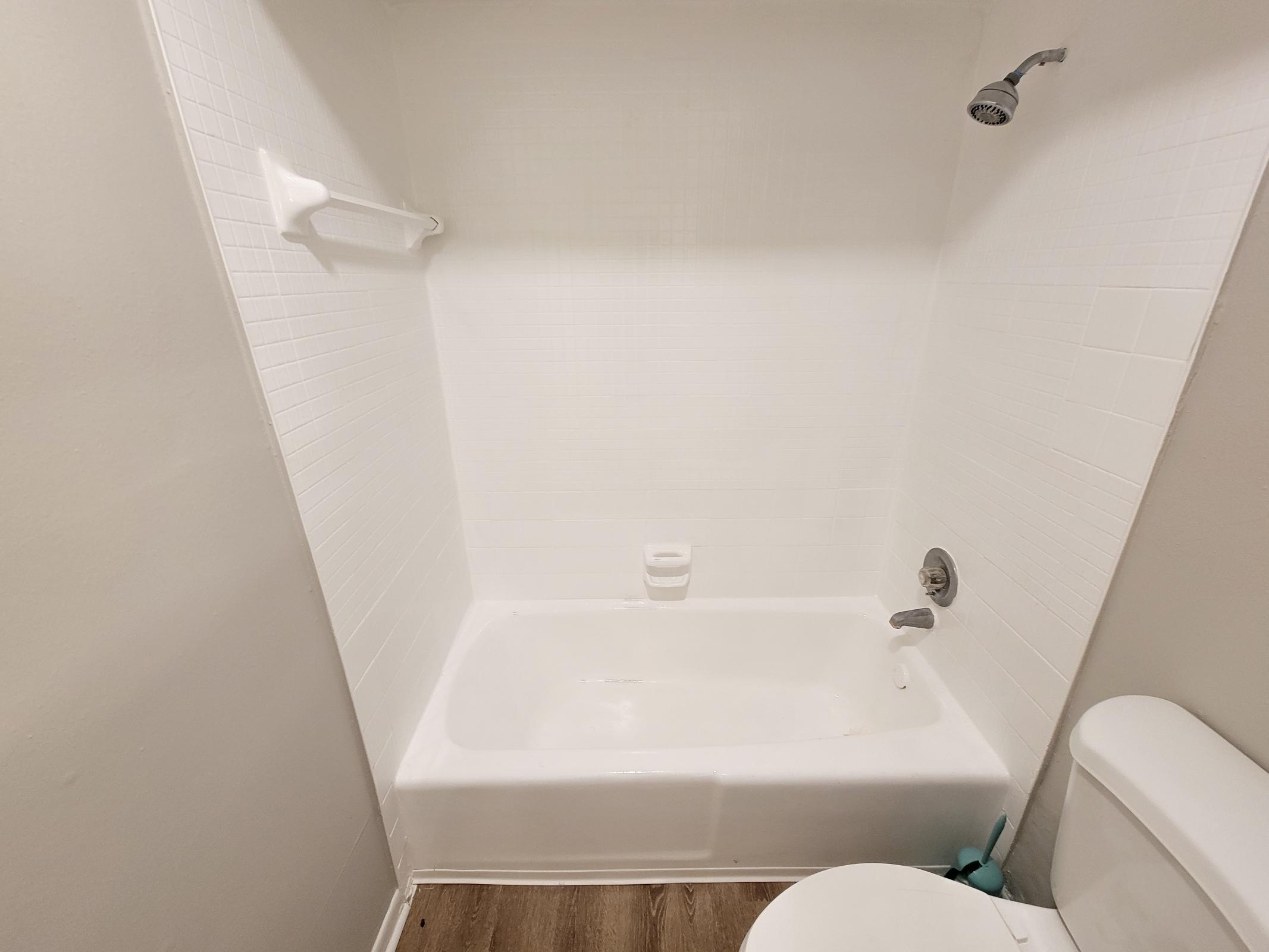 A clean, empty bathroom featuring a white tub with surrounding tiled walls, a showerhead mounted above, and a toilet in the corner. The flooring is wooden, and there is a towel rack on the wall. The space appears bright and minimalistic.