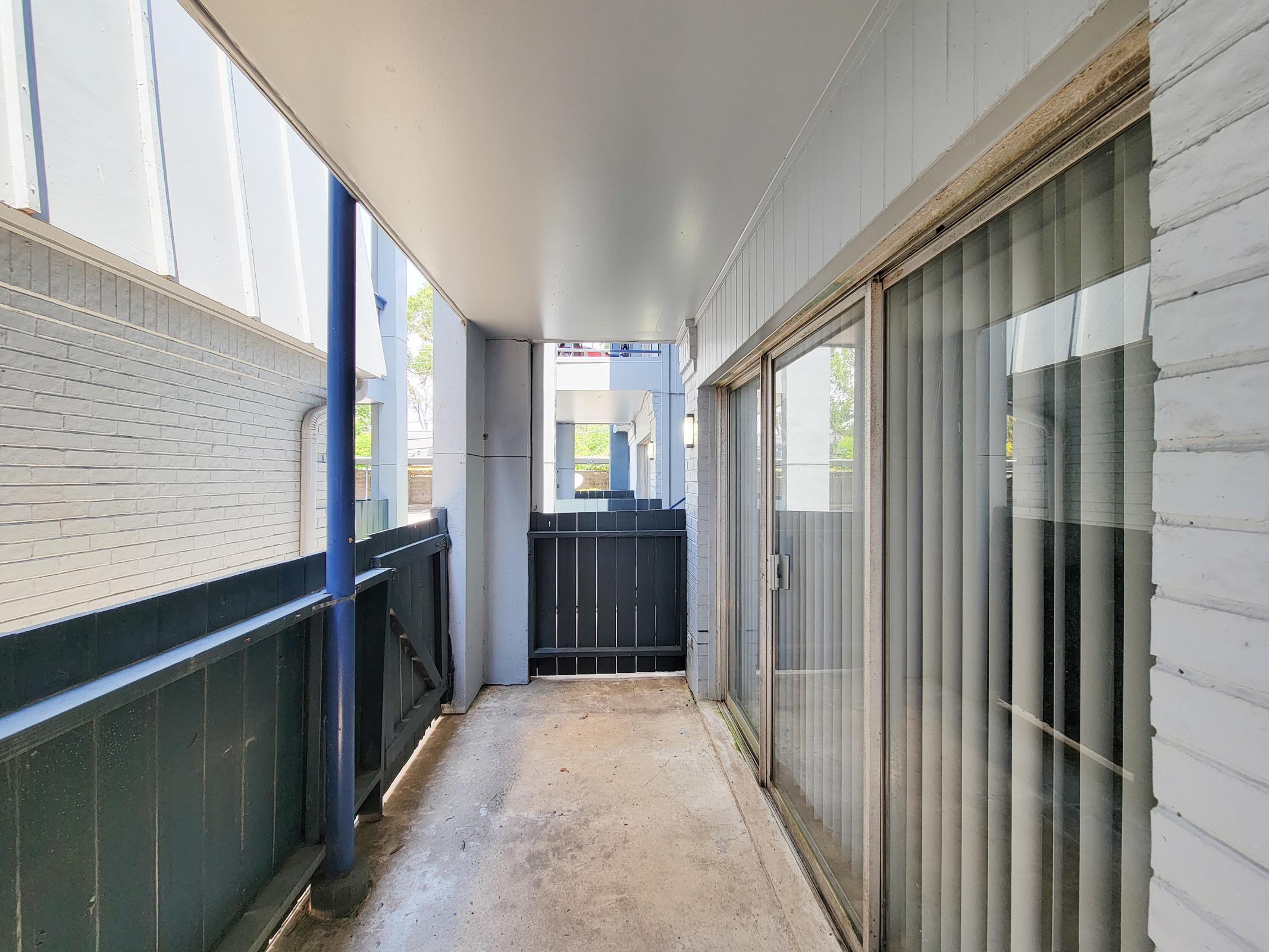 View of a covered balcony or corridor featuring blue and white walls, sliding glass doors, and a concrete floor. Natural light is coming in from the open end, illuminating the space. The area is uncluttered, with a railing visible at the far end.