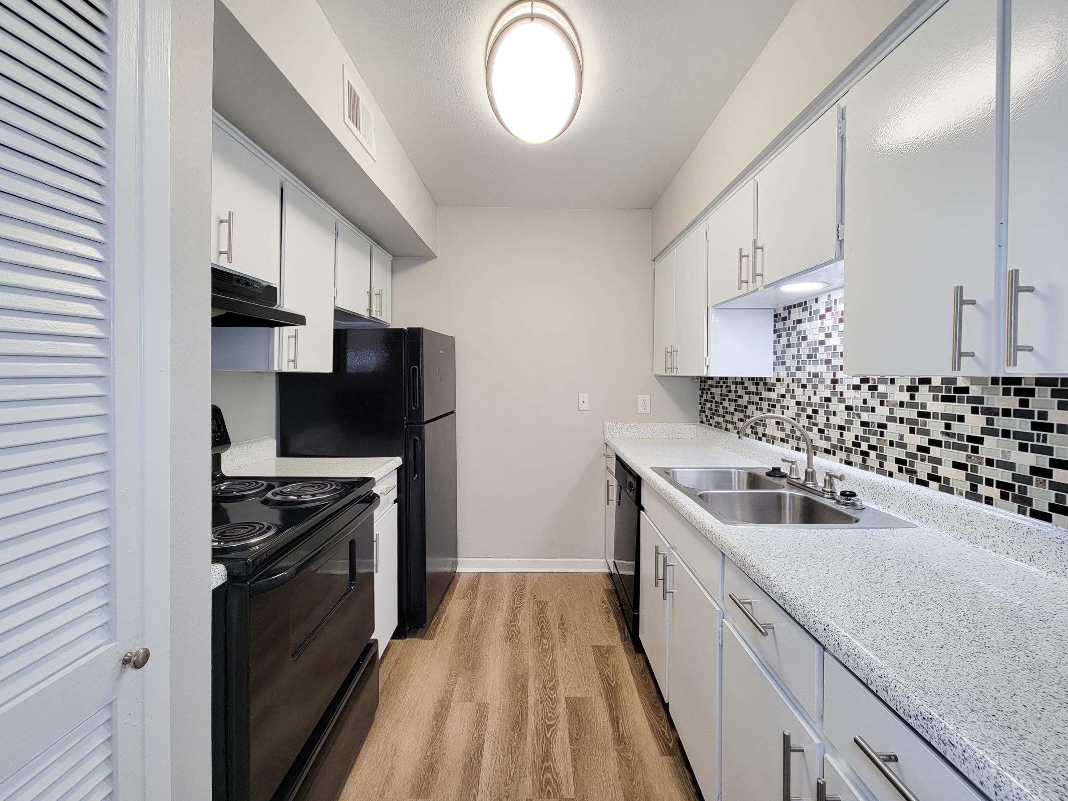 A modern kitchen featuring white cabinets and black appliances. The countertop is light with a speckled design, and the backsplash consists of black and gray tiles. The kitchen has a double sink and a spacious layout with wood-style flooring and a ceiling light fixture.
