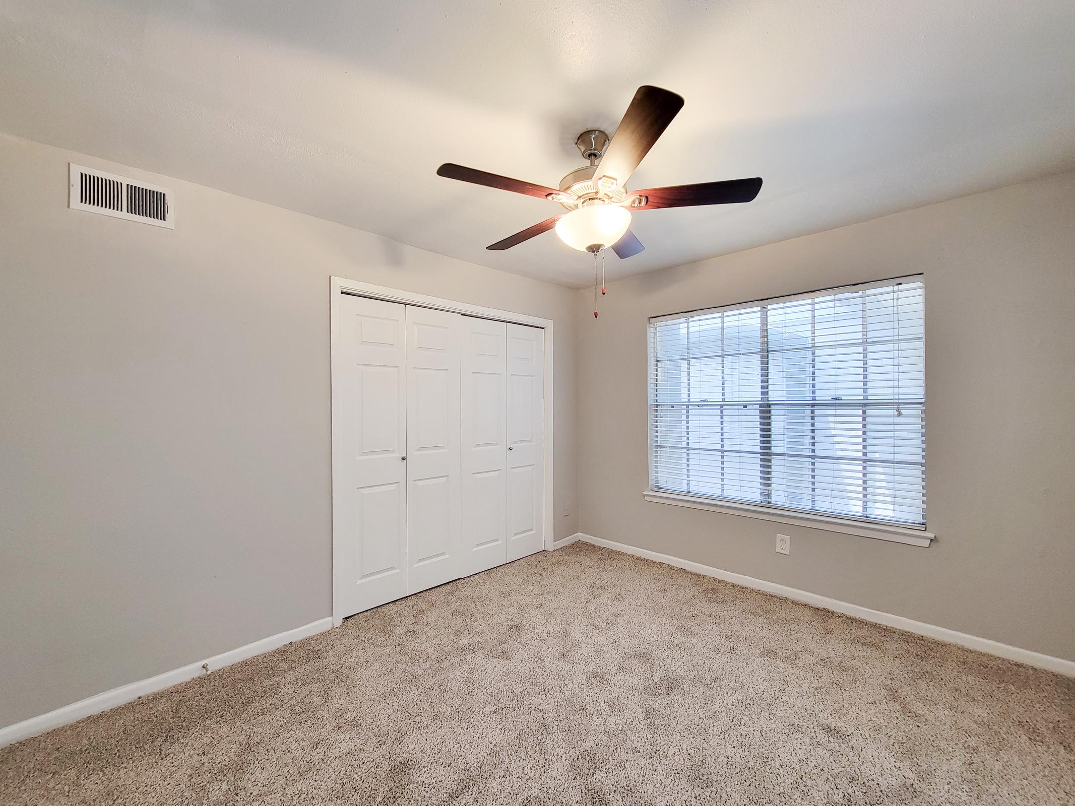 A well-lit room featuring beige walls and carpet, a ceiling fan with light, a window with blinds, and a closet with sliding doors. The overall ambiance is warm and inviting, suitable for various uses such as a bedroom or office space.