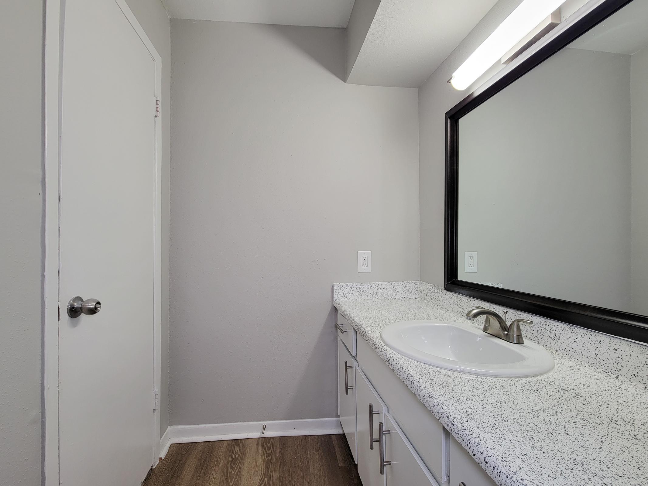 A modern bathroom with light gray walls, a large mirror above a white countertop with a sink, and a door on the left side. The flooring is brown wood-like laminate, and overhead lighting illuminates the space. The overall design is simple and minimalist.