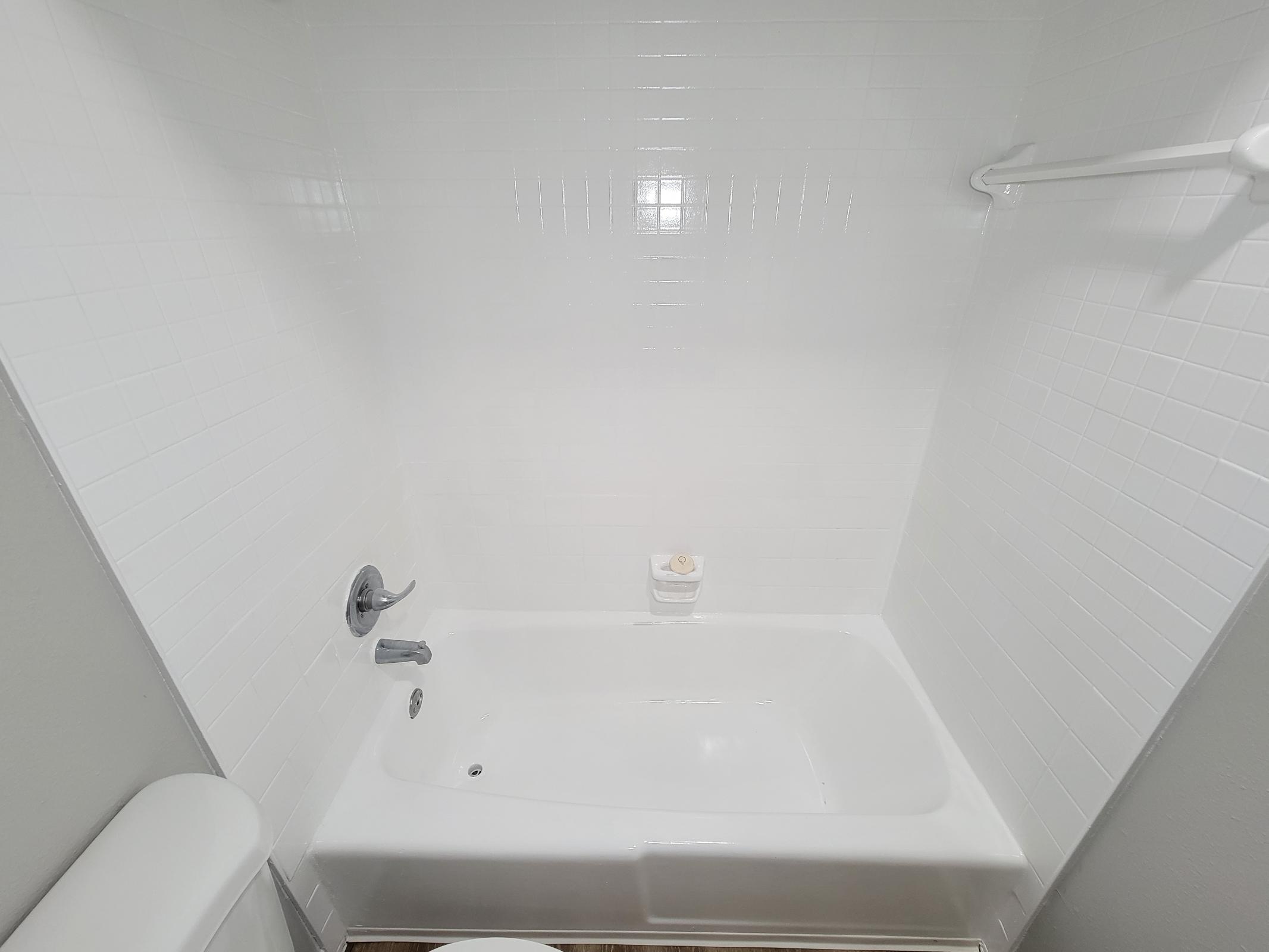 A clean and modern bathroom shower area featuring a white tiled wall and a white bathtub. There is a silver faucet and a soap holder mounted on the wall. The overall appearance is bright and well-maintained, with minimal decor.