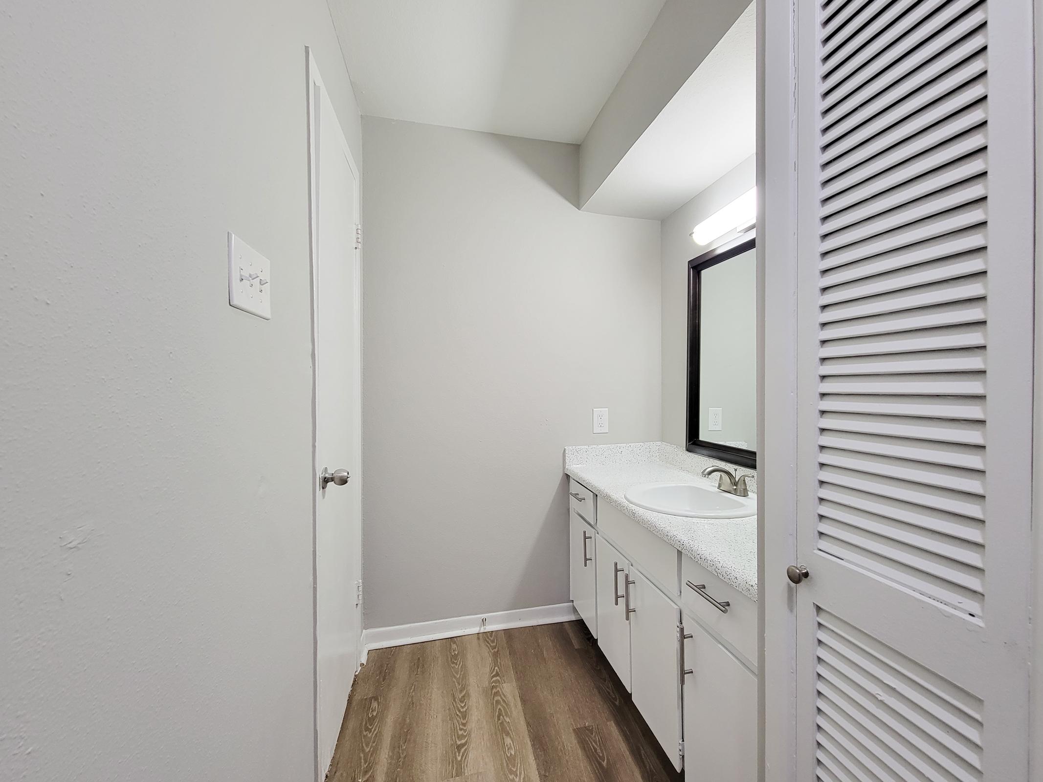 A modern bathroom featuring a light gray color scheme, a double-sink vanity with a white countertop, and a large mirror. The flooring is a wood-like laminate. A door can be seen on the left, and there’s a closed louvered closet door to the right. Bright lighting enhances the clean, spacious appearance.