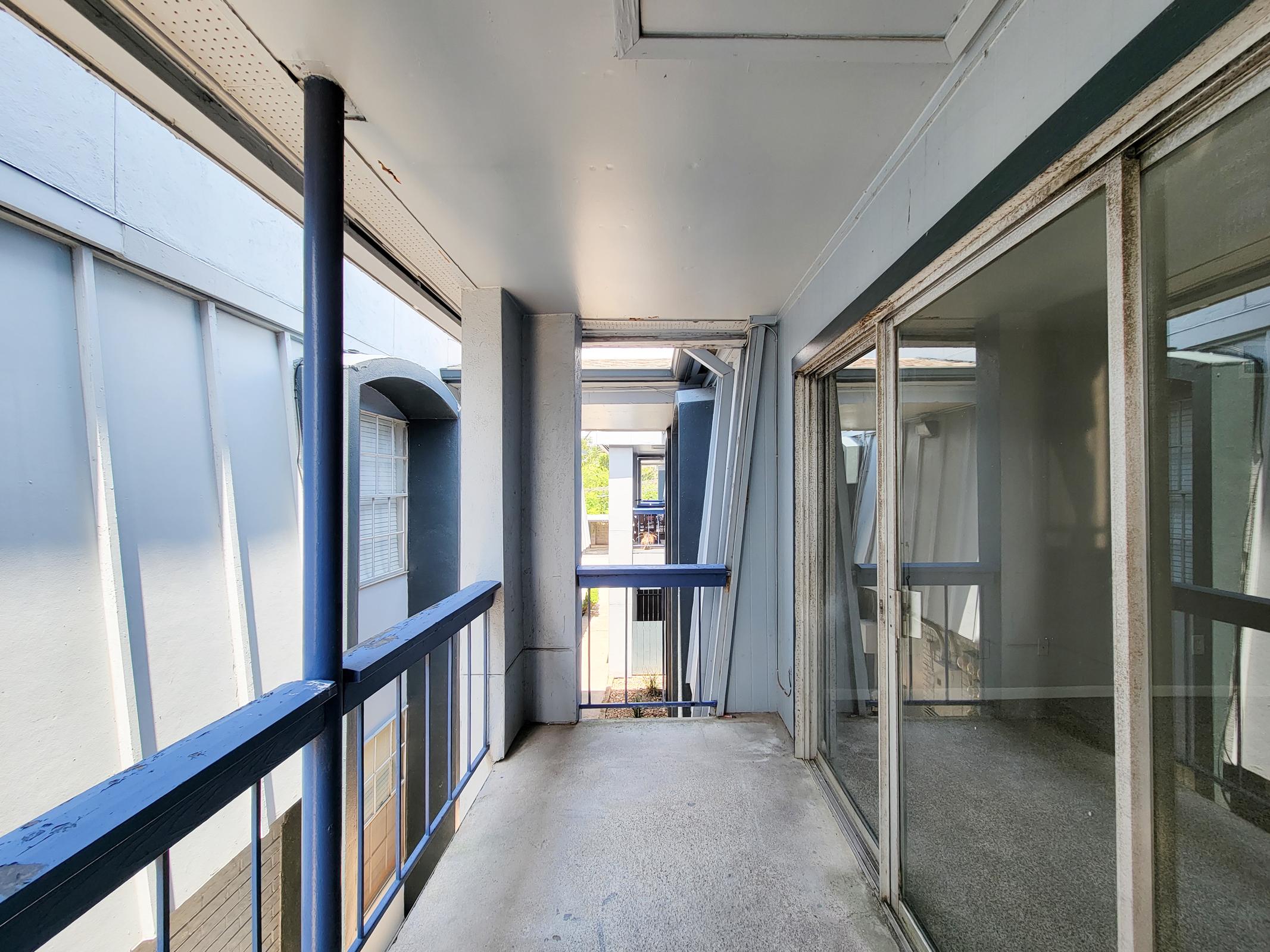 A narrow balcony or corridor with gray walls and a blue railing. On one side, there are large glass windows, and the opposite side features an open space leading to another section of the building. Soft natural light illuminates the area, creating a calm atmosphere.