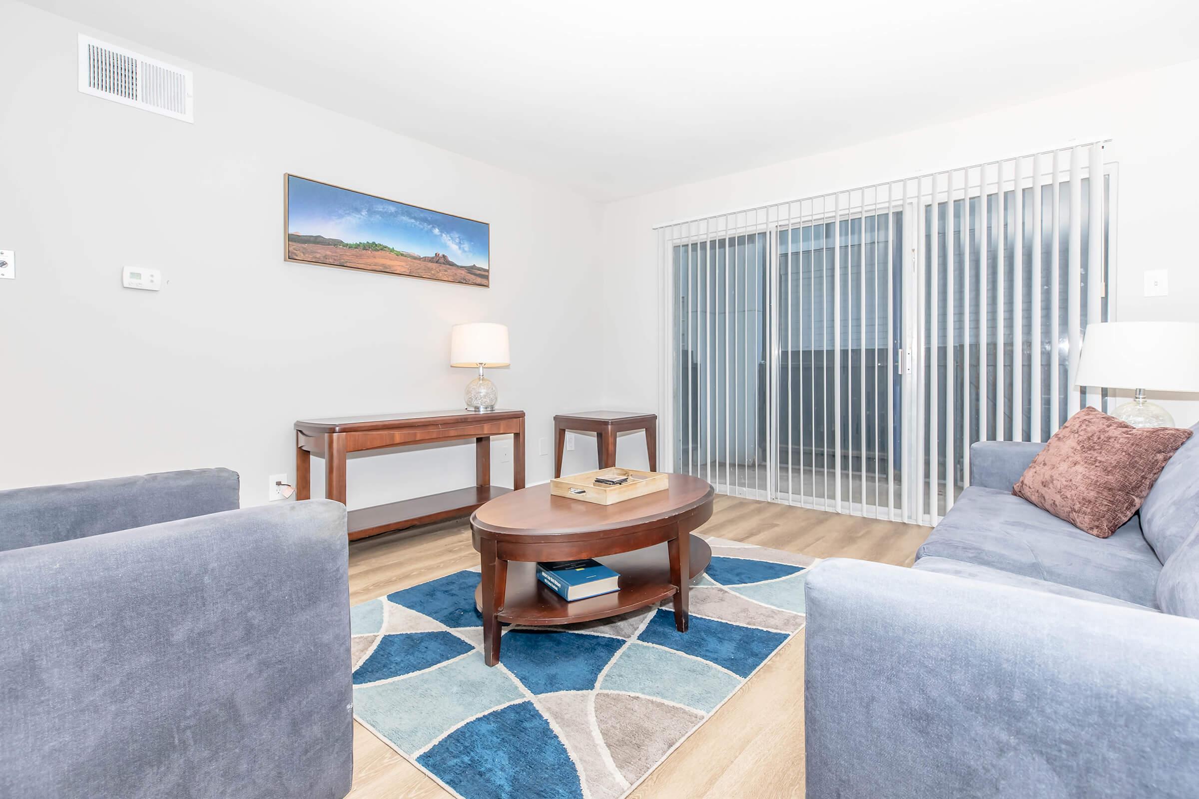 A cozy living room featuring two blue armchairs and a gray sofa, set around a round wooden coffee table on a patterned rug. A lamp sits on a side table, and there's a landscape painting on the wall. Large sliding doors with vertical blinds provide natural light, enhancing the inviting atmosphere.