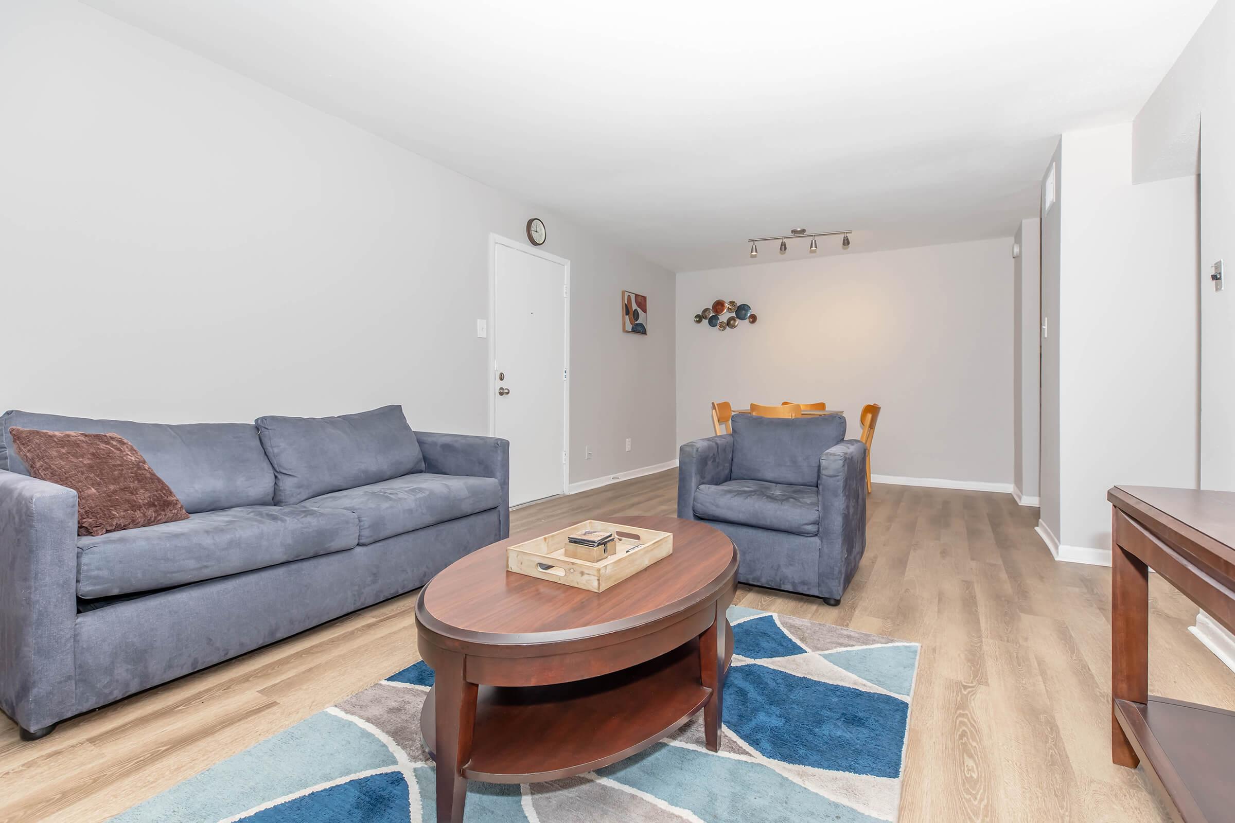 A modern living room with light gray walls, featuring a blue couch, a blue armchair, and a round coffee table. The floor is hardwood, and there is a colorful area rug underneath the table. A dining area is visible in the background with wooden chairs. A clock and artwork are on the walls.