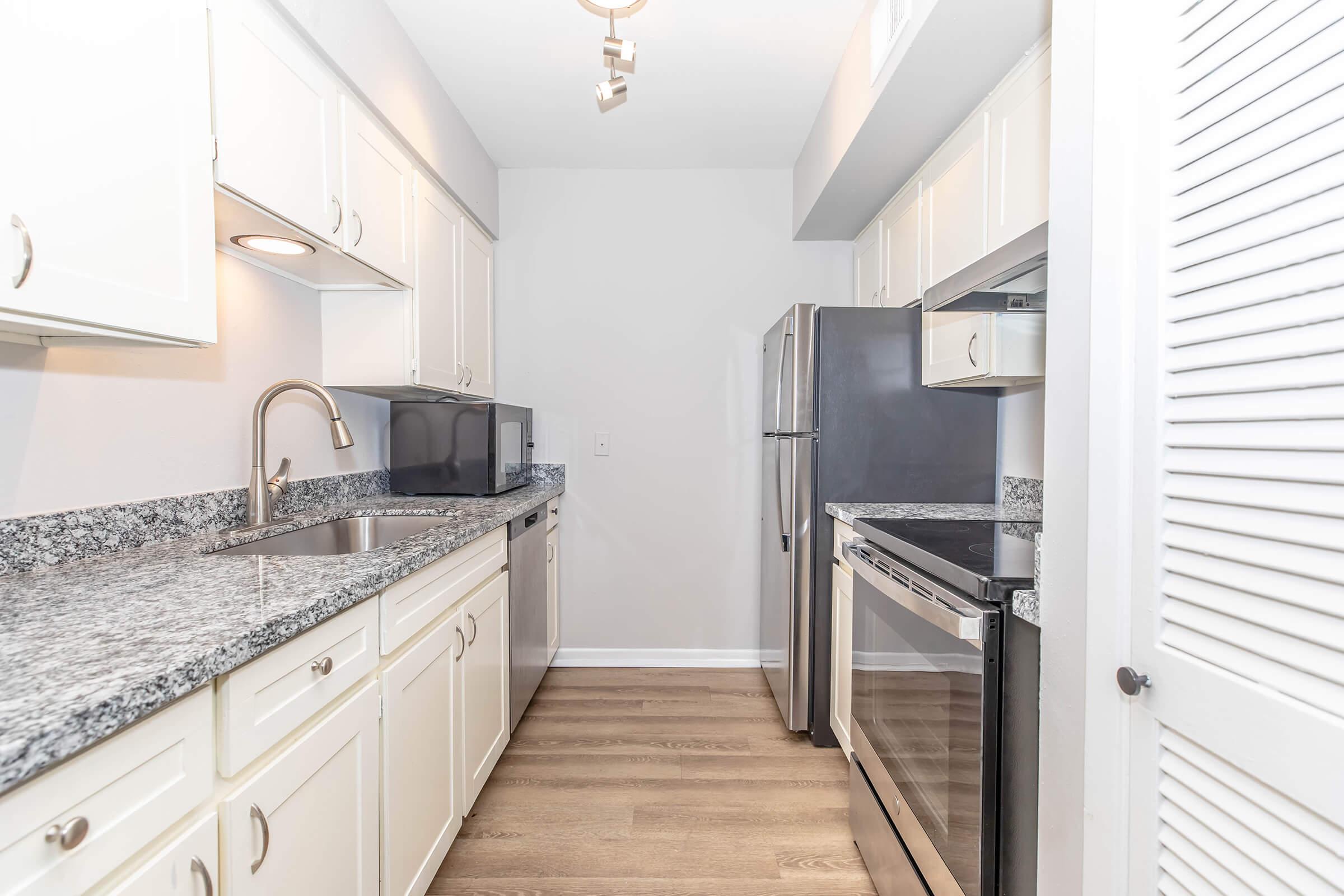 A modern kitchen featuring white cabinets, granite countertops, stainless steel appliances including a refrigerator, stove, and microwave, and a single-basin sink. The space has a neutral wall color and wood flooring, creating a bright and inviting atmosphere.