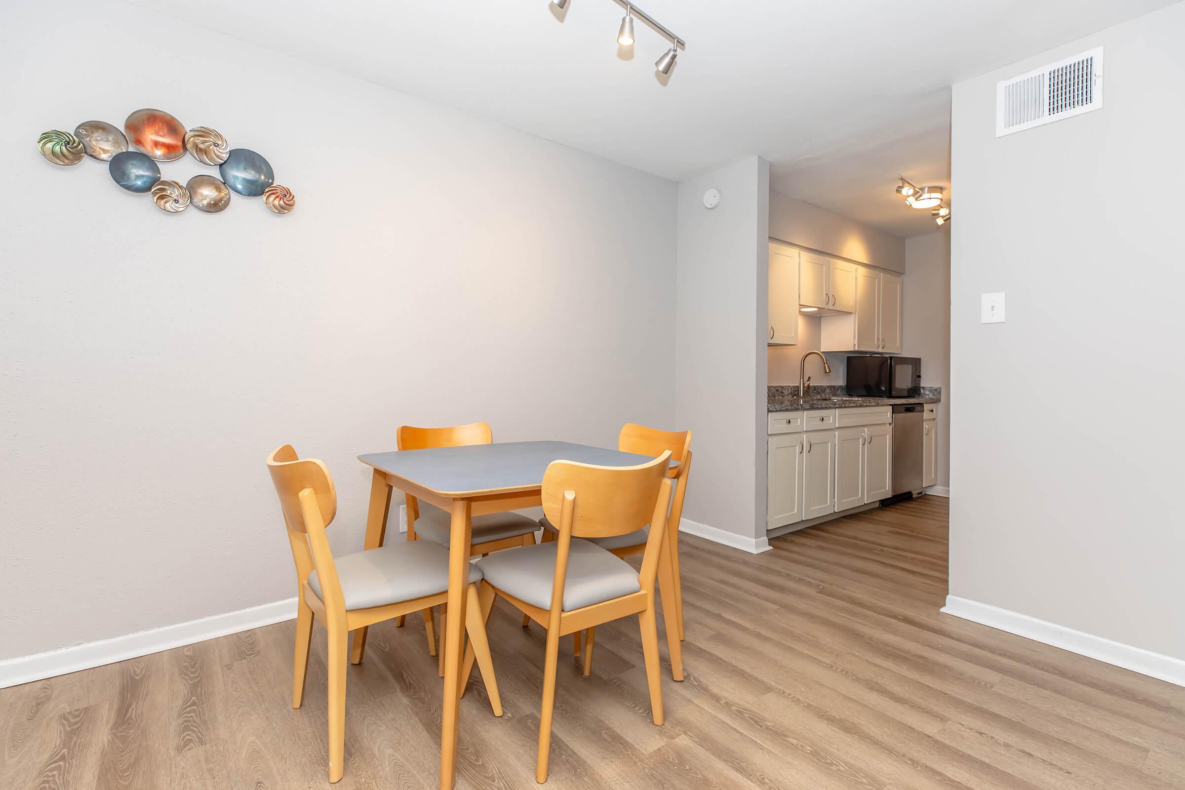 A dining area featuring a small rectangular table with four wooden chairs, set against a light gray wall. Decorative wall art made of colorful circular metal pieces is displayed above the table. In the background, a kitchen area with white cabinetry and appliances is partially visible.