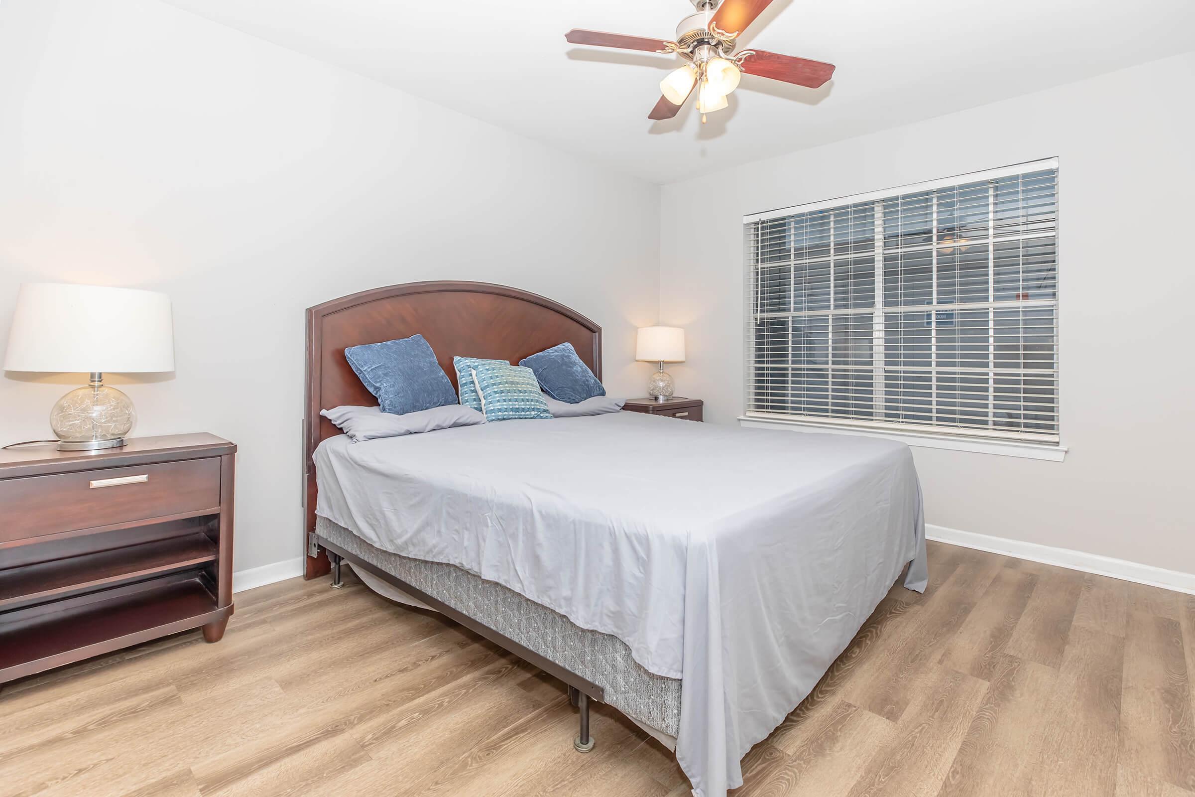 A cozy bedroom featuring a queen-sized bed with a light gray blanket, two decorative pillows, and a wooden headboard. There are two bedside tables with lamps, and a window with blinds allowing natural light to enter. The floor is made of light-colored wood, creating a warm ambiance.