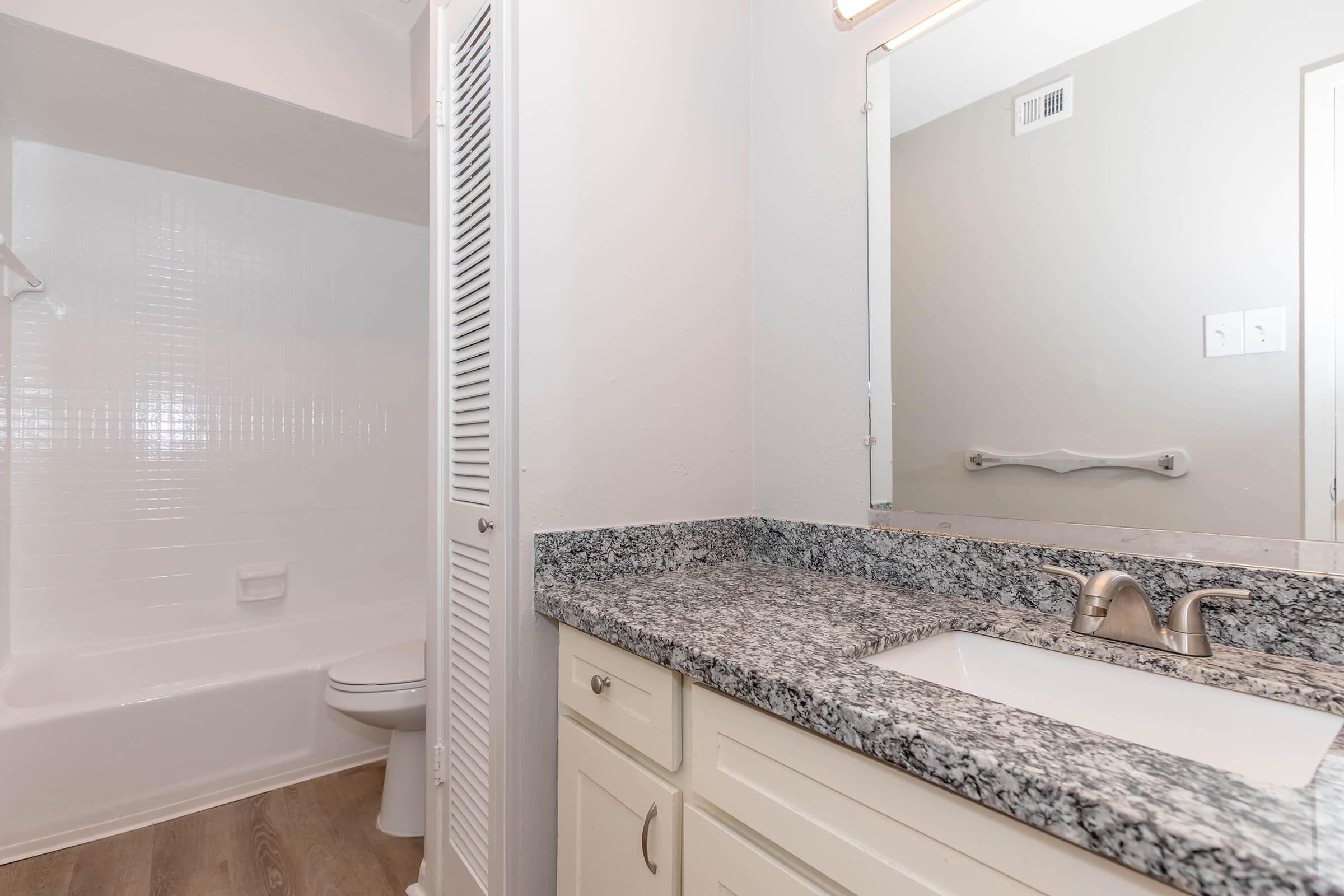 A modern bathroom featuring a granite countertop with a sink, a large mirror, and a white bathtub with a shower. The space has light-colored walls and a white toilet, with wood-like flooring that adds warmth to the room. Bright lighting enhances the clean and spacious appearance.