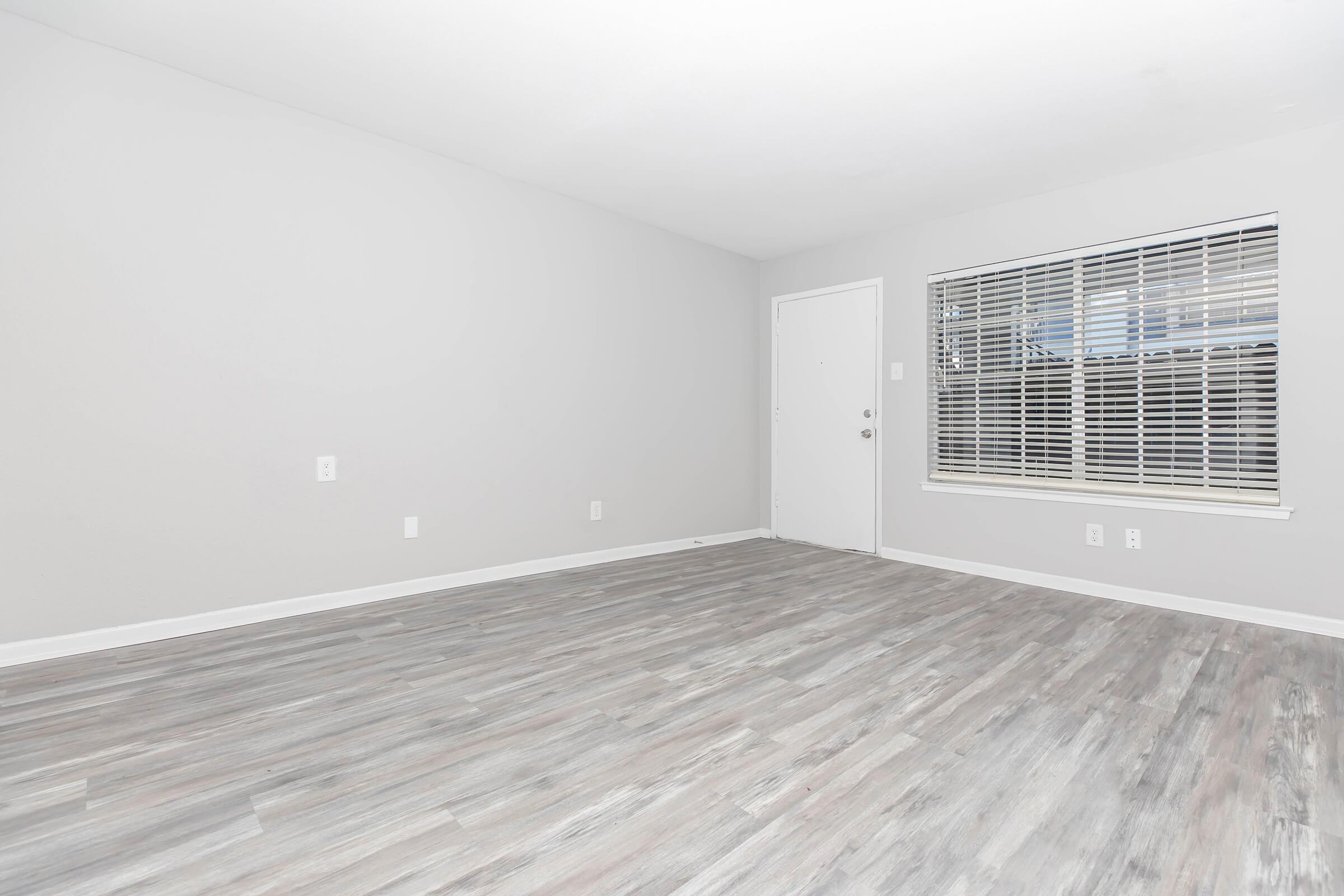 A spacious, empty room featuring light gray walls and wood-like flooring. The room has a large window with blinds, allowing natural light to enter, and a white door on one side. The minimalist design creates an open and airy atmosphere, ideal for various interior decorating options.