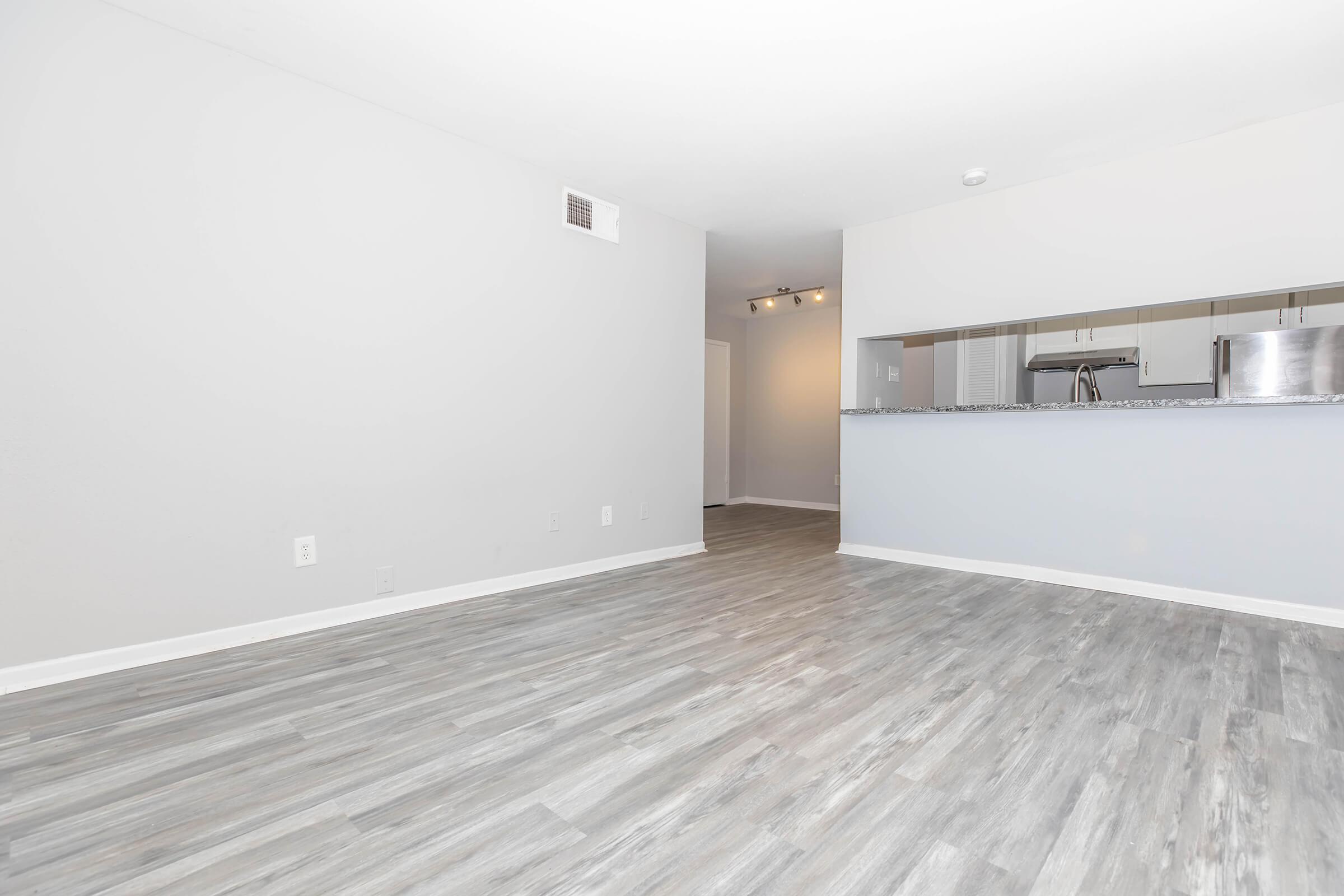 Spacious, empty living area with light gray walls and a wood-style floor. A breakfast bar is visible at the far end, leading to a kitchen area. Natural light illuminates the room, creating a bright and airy atmosphere.