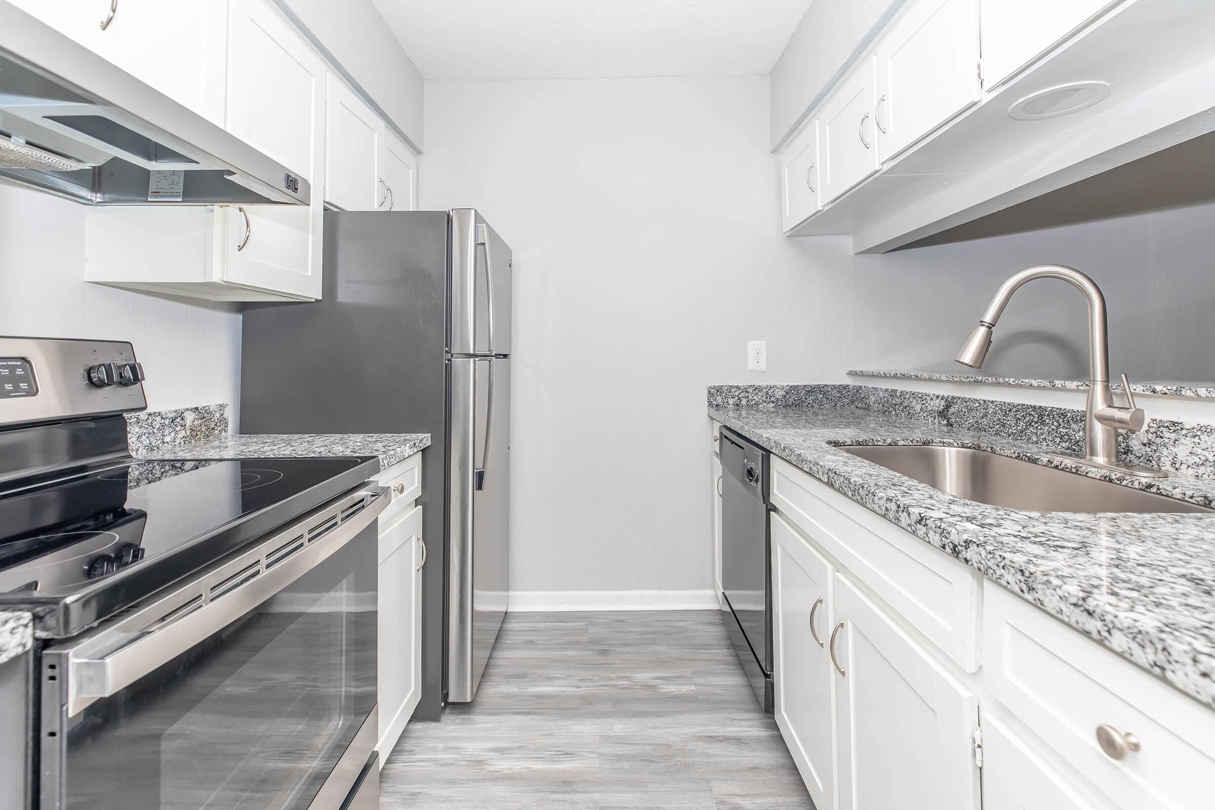 Modern kitchen featuring stainless steel appliances, including a refrigerator, oven, and a sink with a sleek faucet. The countertops are made of speckled granite, and the cabinetry is white, providing a bright and clean appearance. The flooring is light-colored, complementing the overall design.