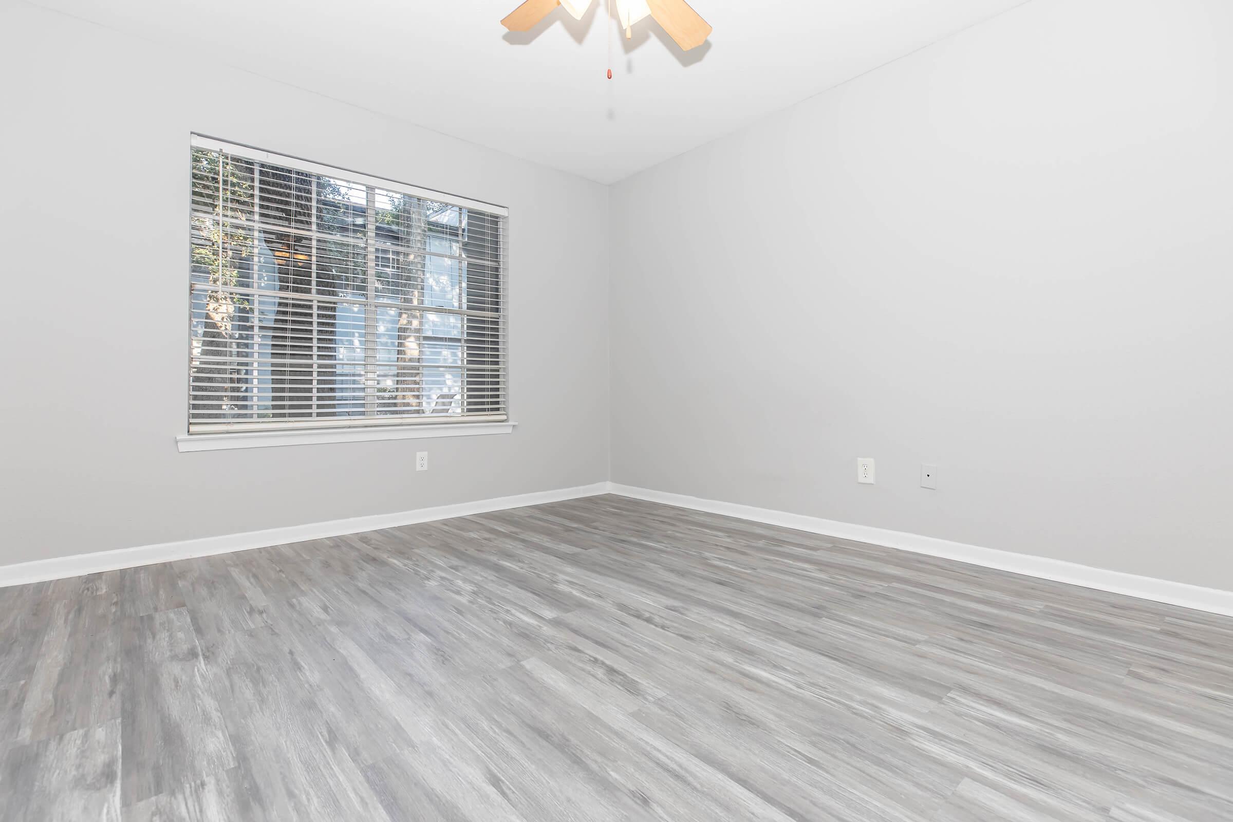 Empty room with light gray walls and a light wood-like floor. A window with horizontal blinds allows natural light to enter, and a ceiling fan is visible. No furniture or decorations are present, creating a minimalist aesthetic.