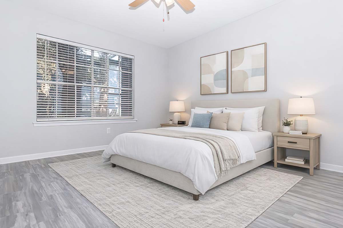 A bright, modern bedroom featuring a large bed with white bedding, two decorative pillows, and a light throw. There are two nightstands with lamps on either side of the bed. The room has a window with white blinds, allowing natural light to enter, and a soft area rug covering the floor. Abstract artwork hangs on the walls.