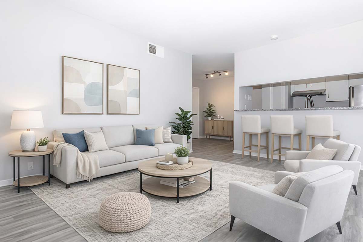 A modern, minimalist living room featuring a light-colored sofa with decorative pillows, a round coffee table, and a textured area rug. There are two armchairs, a floor lamp, and indoor plants. The room is bright with soft wall art and an open kitchen area visible in the background.