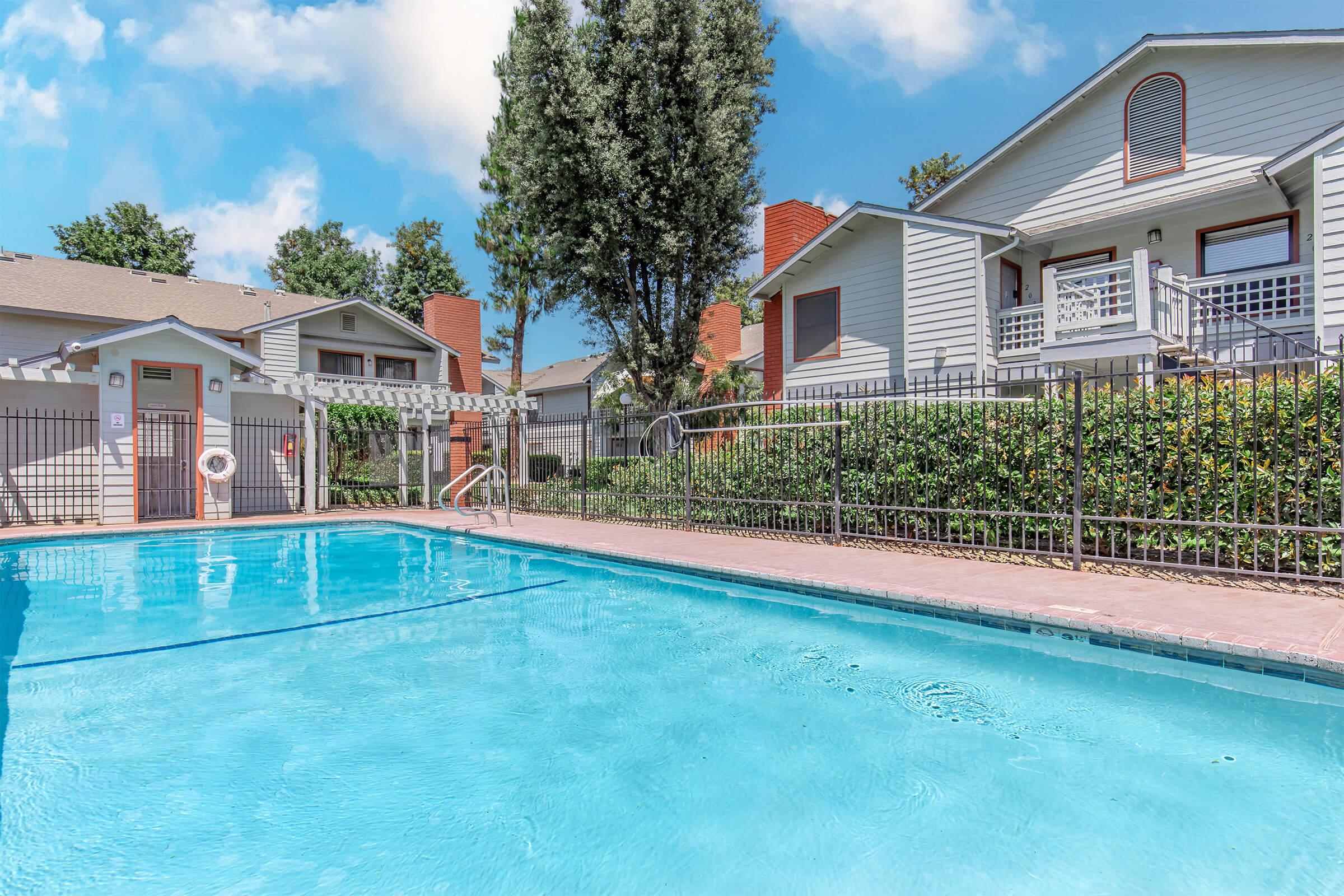 A clear blue swimming pool surrounded by a fence, with residential buildings in the background. Lush greenery lines the sides, and a few trees are visible. The sky is bright with scattered clouds, creating a serene atmosphere.