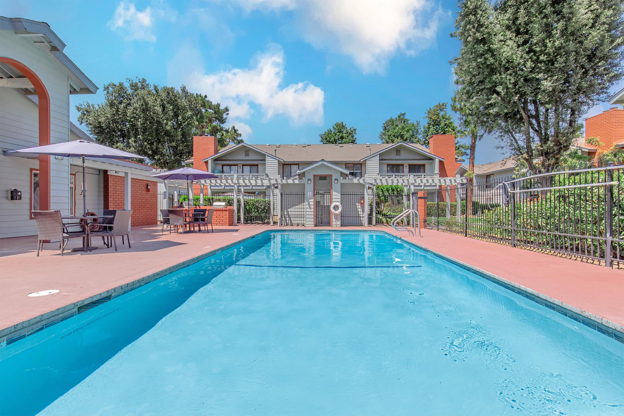 A sunny view of a pool area with clear blue water surrounded by a fenced patio. In the background, there are a couple of residential buildings, trees, and outdoor seating with umbrellas, creating a relaxing atmosphere. The sky is bright with fluffy white clouds.