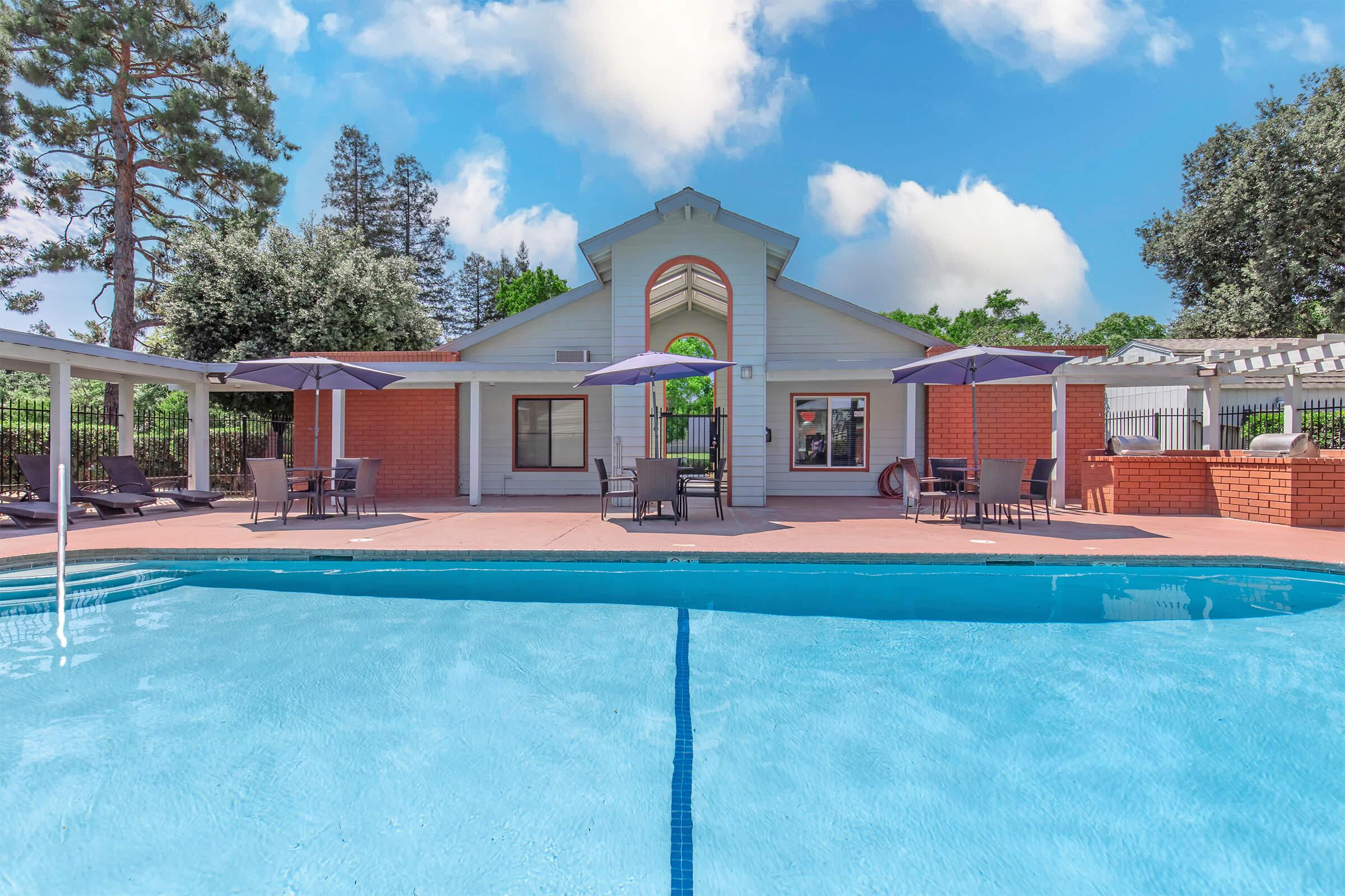 A clear blue swimming pool in the foreground with lounge chairs and umbrellas surrounding it, leading up to a charming house. The house features large windows and a peaked roof, situated in a lush, green environment under a partly cloudy sky.