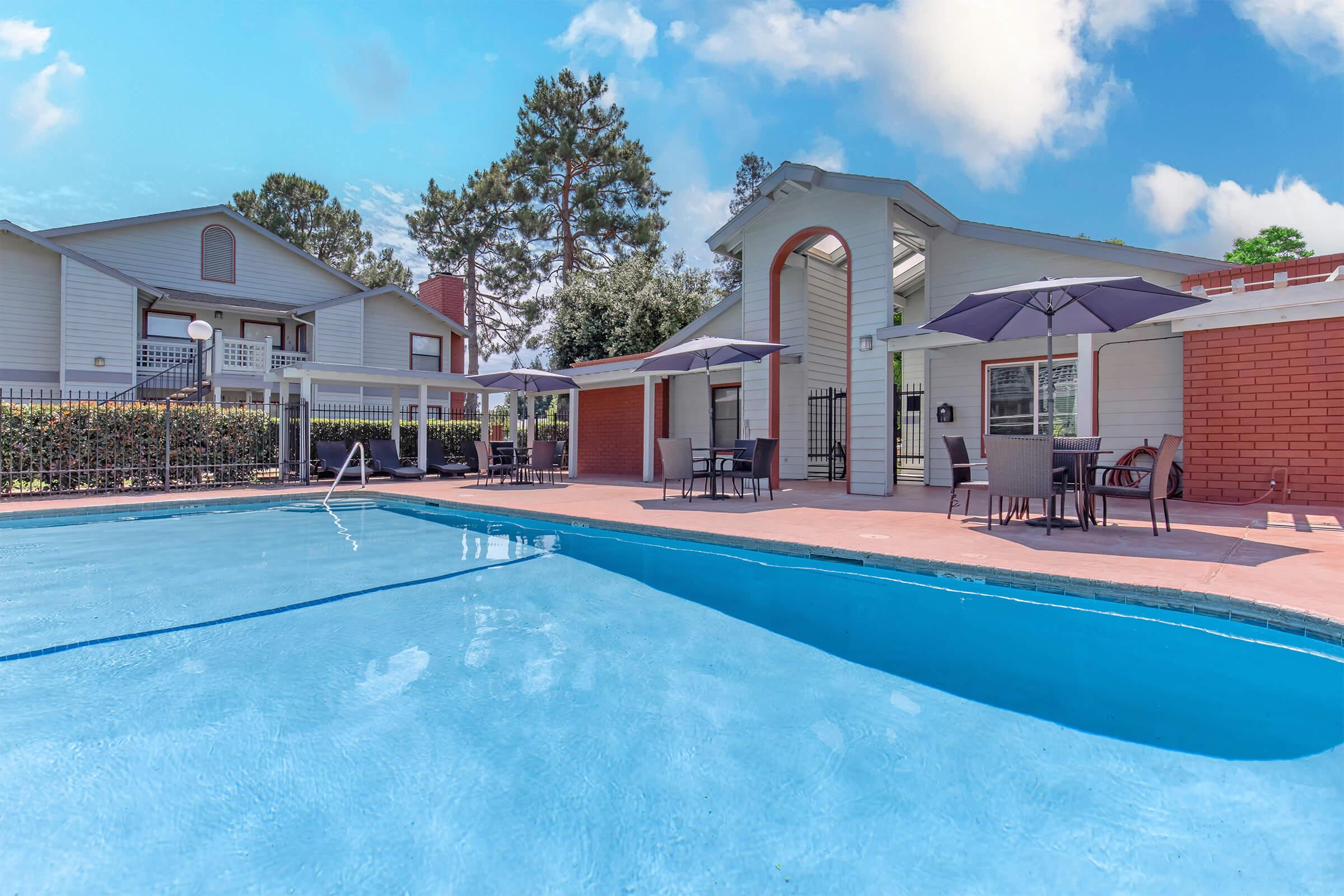 A clear blue swimming pool surrounded by a paved area with lounge chairs and umbrellas. In the background, there are two residential buildings with light-colored siding and red brick accents, along with green trees and a blue sky with fluffy clouds.