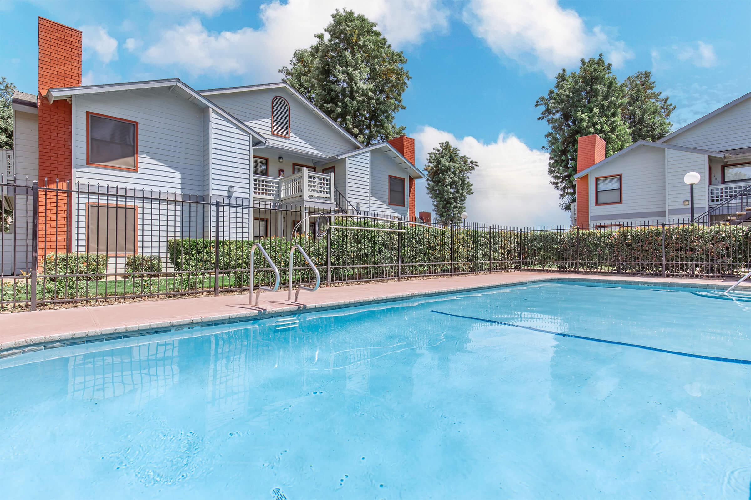 A clear blue swimming pool with a ladder, surrounded by a black metal fence. In the background, there are two light blue apartment buildings with balconies. Lush green trees and bushes are present around the area, under a bright blue sky with a few clouds.