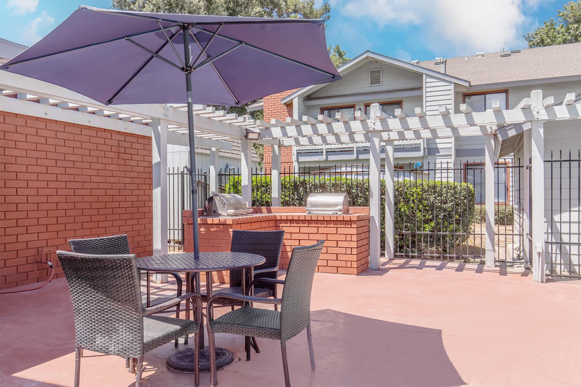 A patio area featuring a round table with four chairs, an umbrella for shade, and a built-in grill surrounded by a brick wall. Lush greenery is visible in the background along with a fence and a two-story building with multiple windows. The sky is partly cloudy.