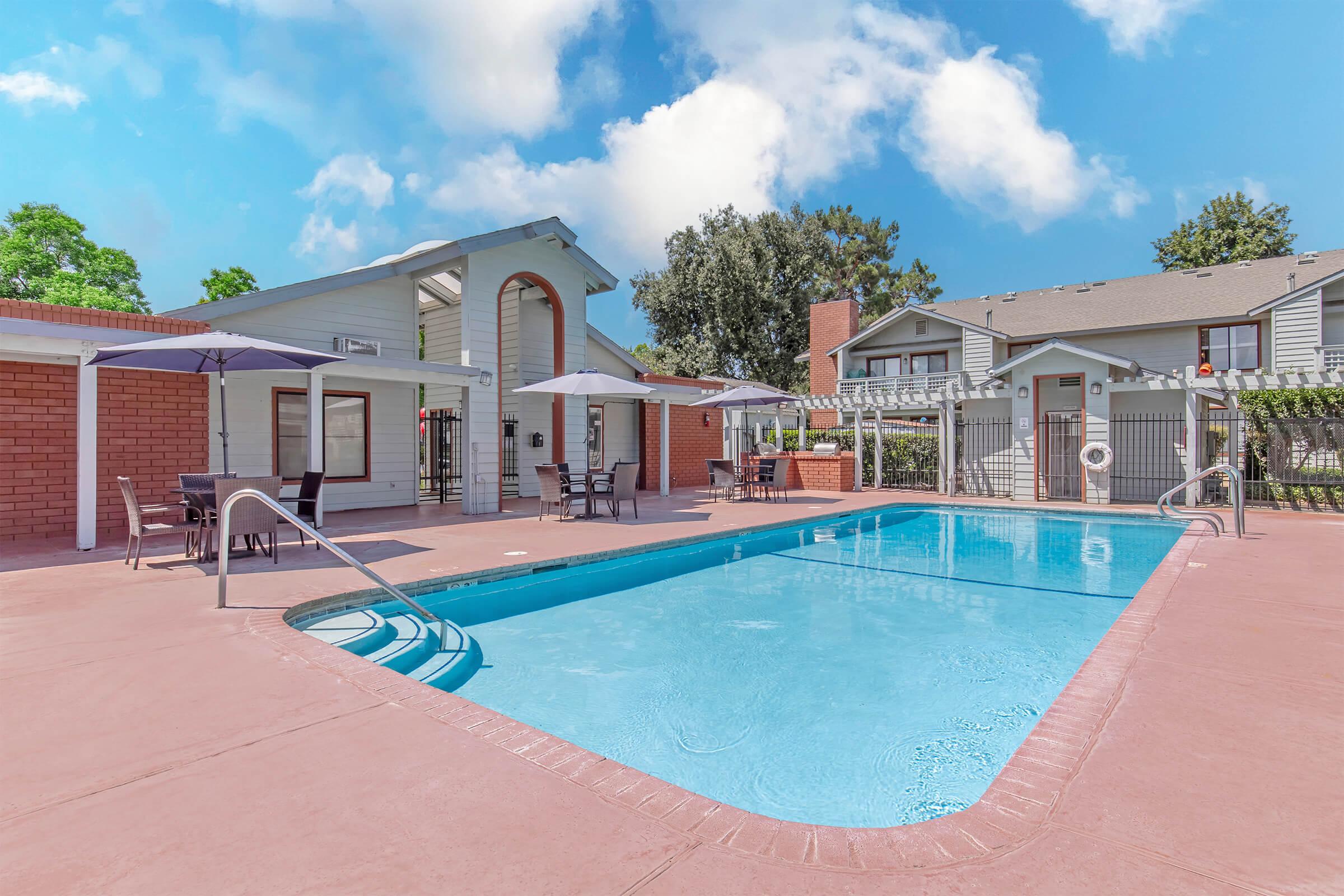 A bright and inviting outdoor swimming pool area featuring a clear blue pool, surrounded by patio spaces with tables and umbrellas. Lush greenery and residential buildings are visible in the background under a partly cloudy sky. The scene conveys a relaxed and enjoyable atmosphere.