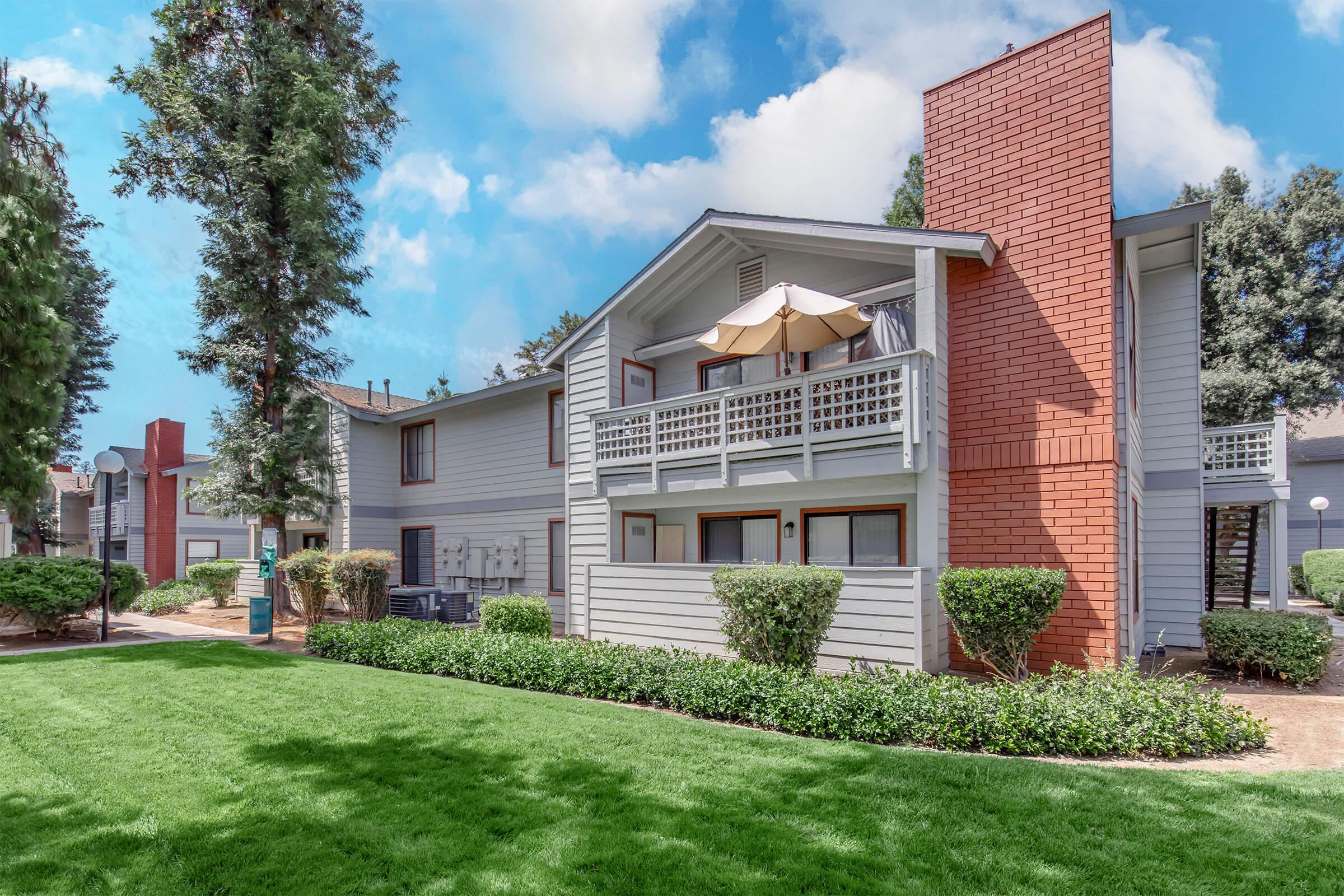 A well-maintained apartment complex with two-story buildings adorned with balconies. The property features green lawns, shrubs, and trees under a blue sky with clouds. A patio umbrella is visible on one of the balconies, creating a welcoming outdoor space.