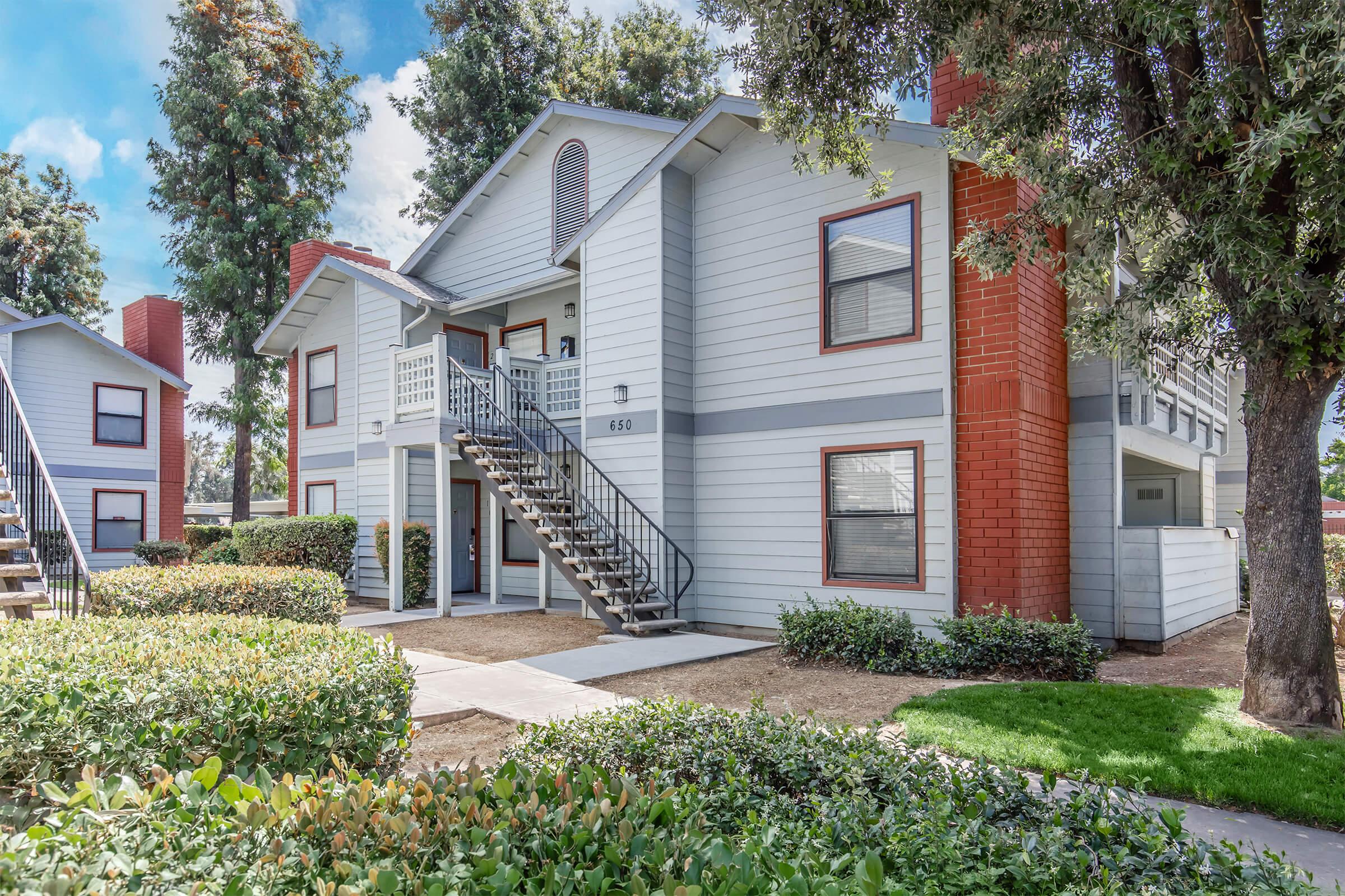 Two-story apartment building with a light blue exterior and red brick accents. Stairs lead to the entrance of the upper level. Lush green landscaping with shrubs and trees surrounds the building. Clear blue sky with scattered clouds in the background.