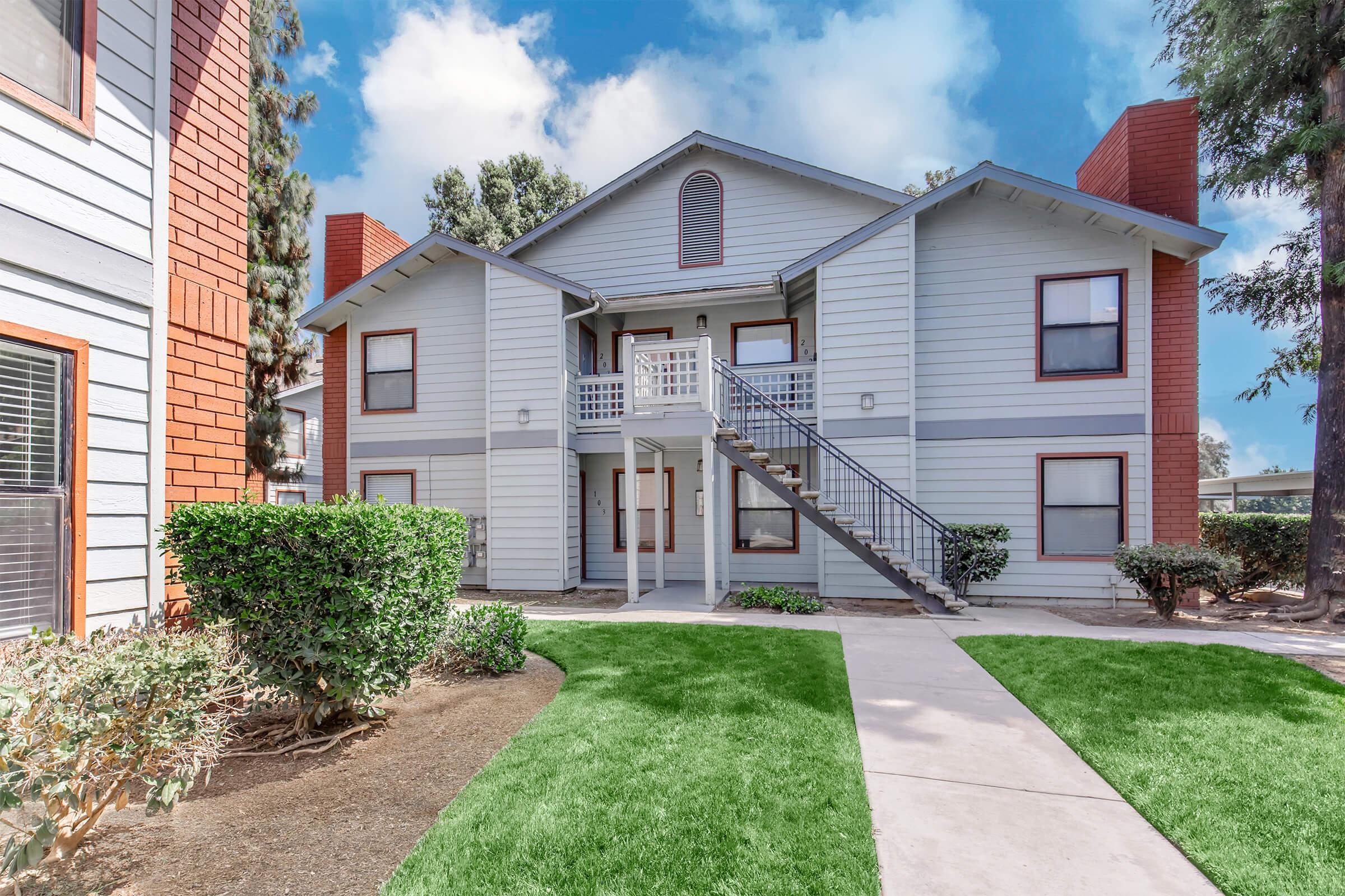 A two-story apartment building with a light blue exterior, featuring red brick accents. There are stairs leading to the upper level, and well-maintained greenery surrounds the building. The sky above is partly cloudy, adding a bright atmosphere to the scene.