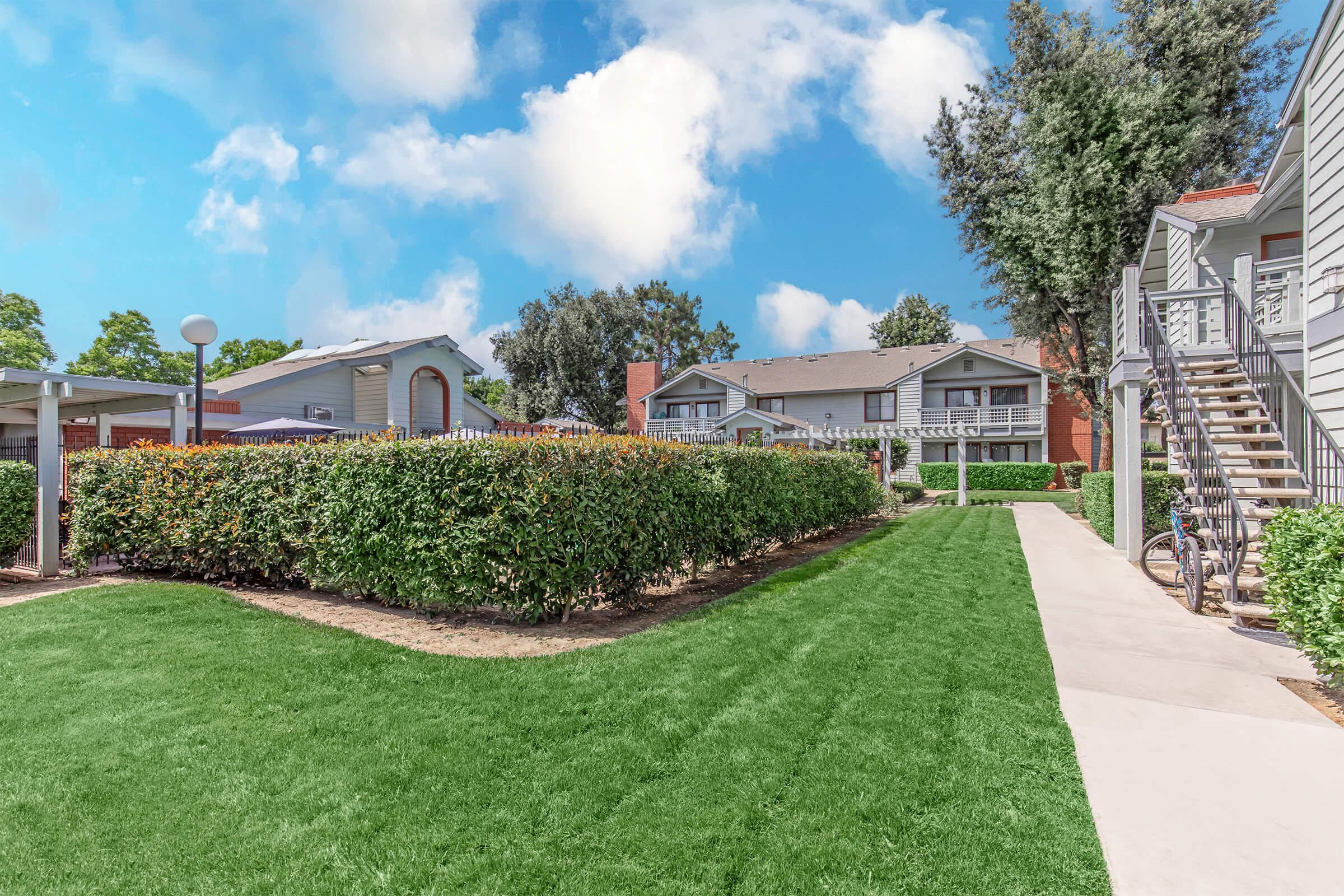 A landscaped outdoor space featuring well-maintained grassy areas, bordered by hedges. In the background, multiple light gray residential buildings with staircases are visible, along with a bike parked on the path. The sky is bright blue with fluffy white clouds, creating a cheerful, inviting atmosphere.