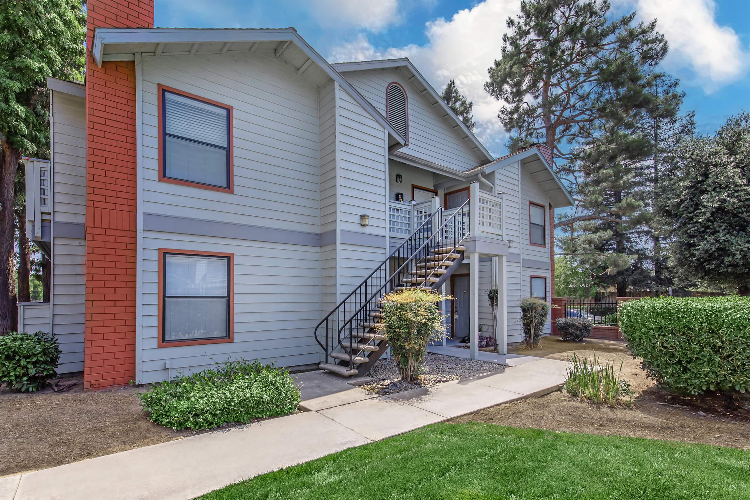 A two-story residential building with a light blue exterior and orange accents. It features a set of exterior stairs leading to the second floor, a small porch area, and well-maintained landscaping including shrubs and grass. Trees are visible in the background under a partly cloudy sky.