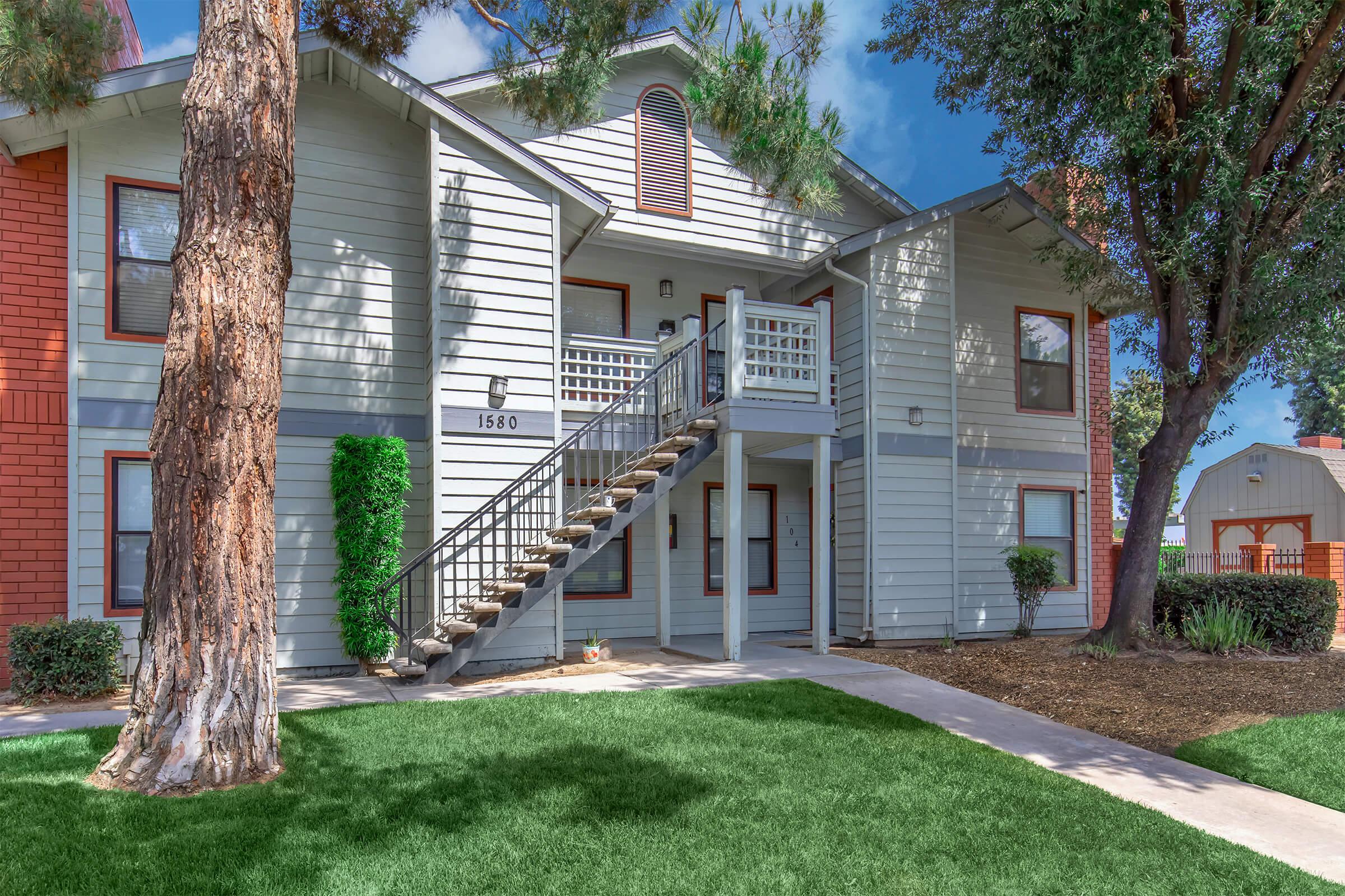 Exterior view of a two-story apartment building featuring a wooden staircase leading to the upper level. The building is surrounded by green grass and landscaping, with trees providing shade. The structure has a light gray color with red accents on some windows. A path leads to the entrance, creating a welcoming atmosphere.