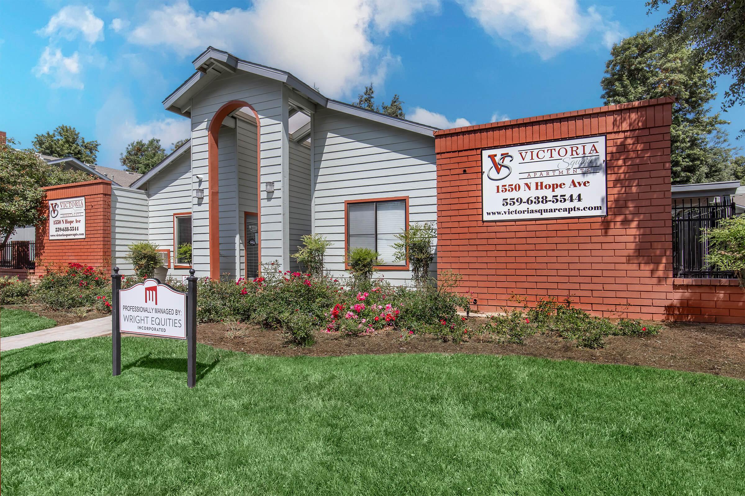 Exterior view of a multi-unit residential building with a modern design. The façade features a mix of gray and orange brick, with signs displaying "Victoria" and contact information. The well-maintained landscaping includes bushes and flowers, along with a sign for "Victoria Managed by Wright Equities."
