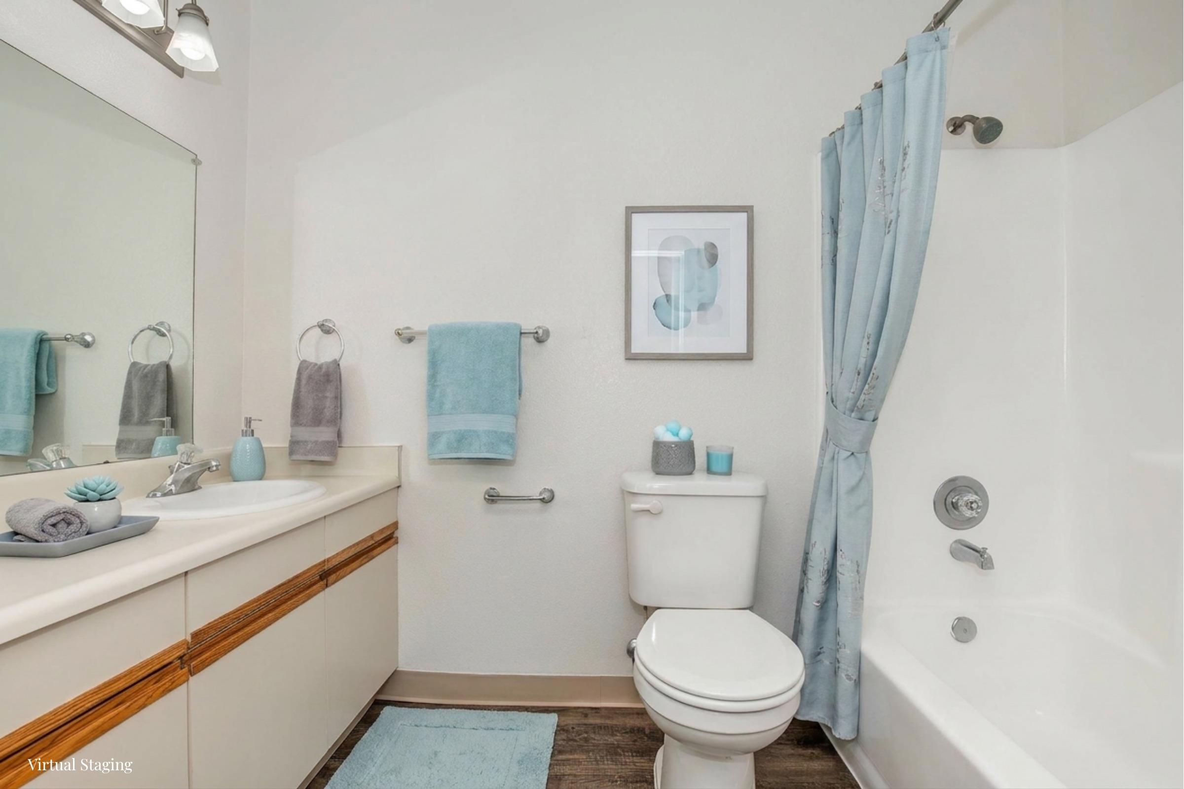 A clean and modern bathroom featuring a bathtub, toilet, and sink. The decor includes light blue towels and accessories, a framed wall art piece, and a neutral color scheme. The lighting is bright, and the overall design is simple and functional.