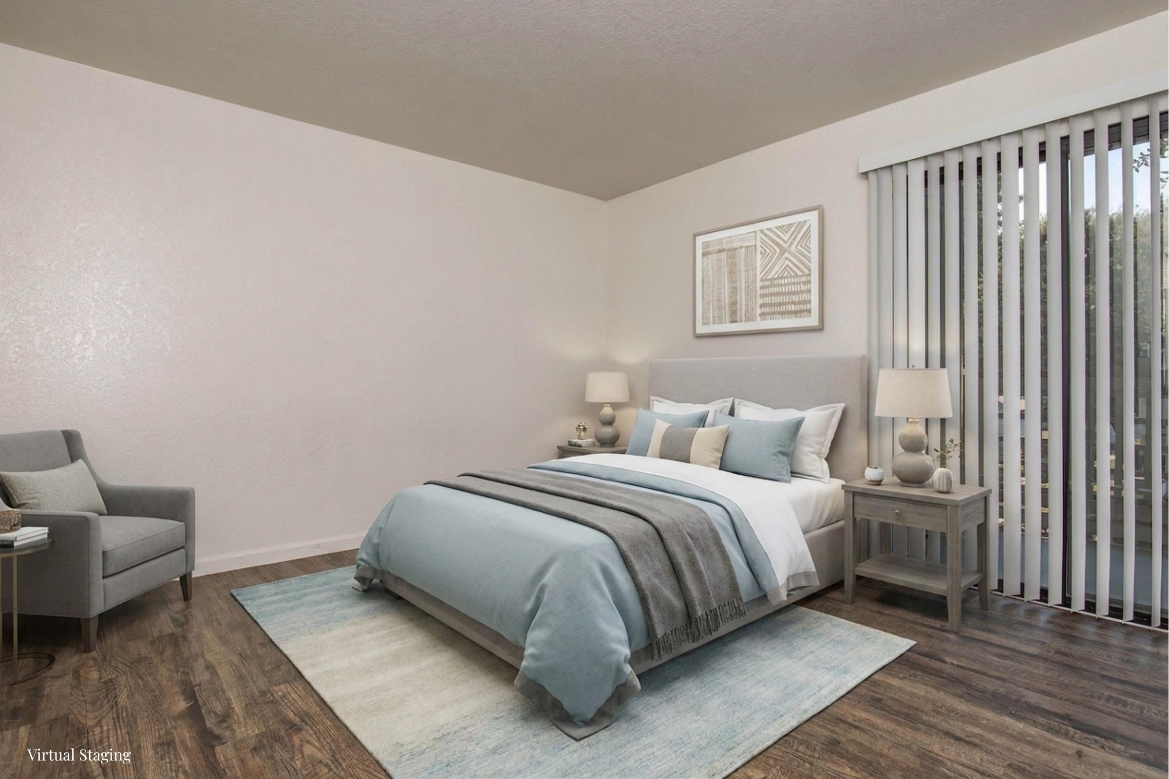 A cozy, well-decorated bedroom featuring a light gray bed with soft blue and white bedding. There are two bedside tables with lamps, a comfortable armchair in the corner, and large sliding doors leading to a patio area. A decorative artwork hangs above the bed, and a soft area rug is placed on the wooden floor.