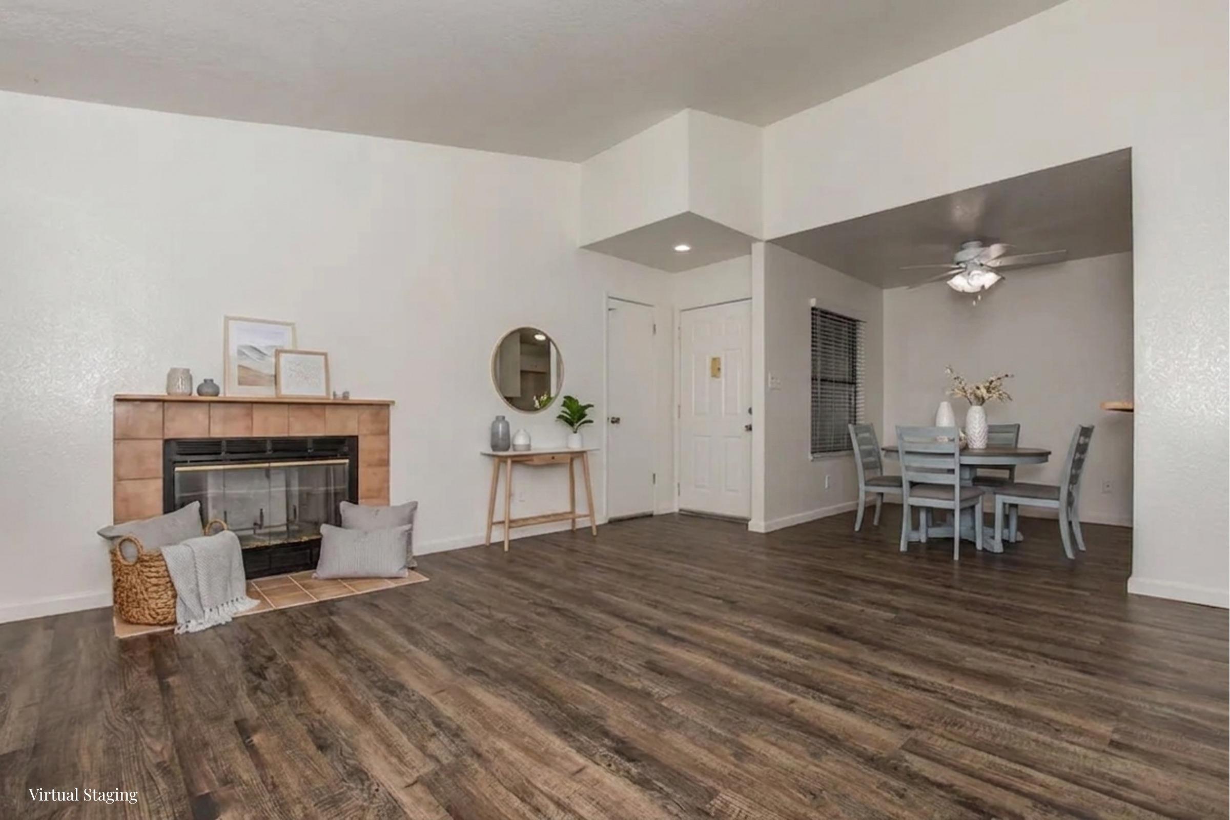 A modern living space featuring a cozy fireplace with decorative pillows and a woven basket. Light-colored walls complement the dark wooden flooring. To the right, a dining area includes a light wooden table with chairs and a ceiling fan. A round mirror and decor on the mantle add a touch of style.