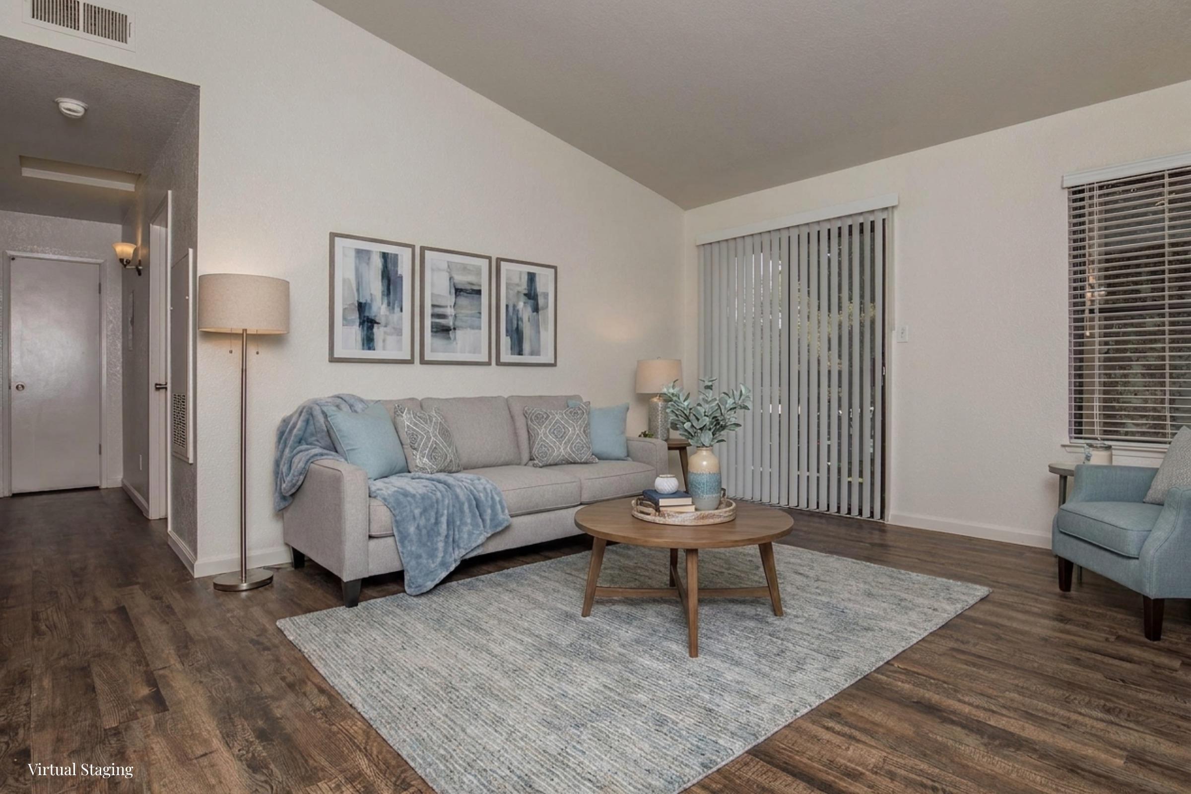A cozy living room featuring a light gray sofa adorned with blue throw pillows, a round wooden coffee table with decorative items, and a soft area rug. The room has light-colored walls, framed abstract art on the wall, a floor lamp, and vertical blinds covering a large window, creating a warm and inviting atmosphere.
