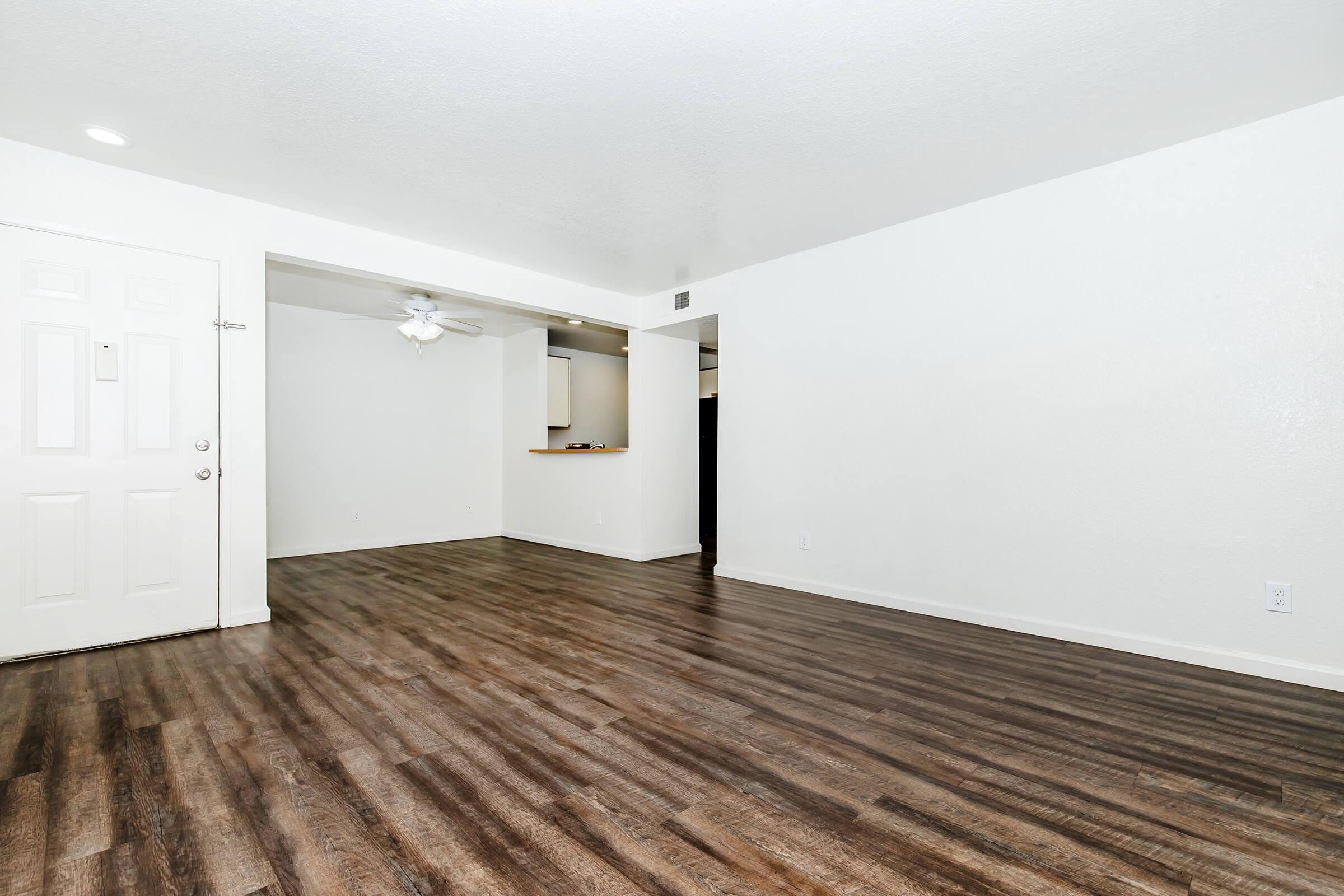A spacious interior view of an apartment featuring light-colored walls and a wooden floor. The image shows an empty living area with a ceiling fan and a doorway leading to another room. Natural light is present, enhancing the bright and airy feel of the space.