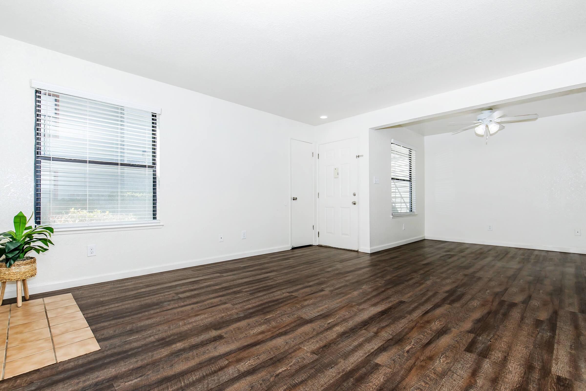 Bright, spacious living area with white walls and large windows. Features a light wood-like floor and a small potted plant in the corner. The room is open and airy, with a ceiling fan visible in the adjoining area. Doorways lead to other rooms, creating a welcoming atmosphere.
