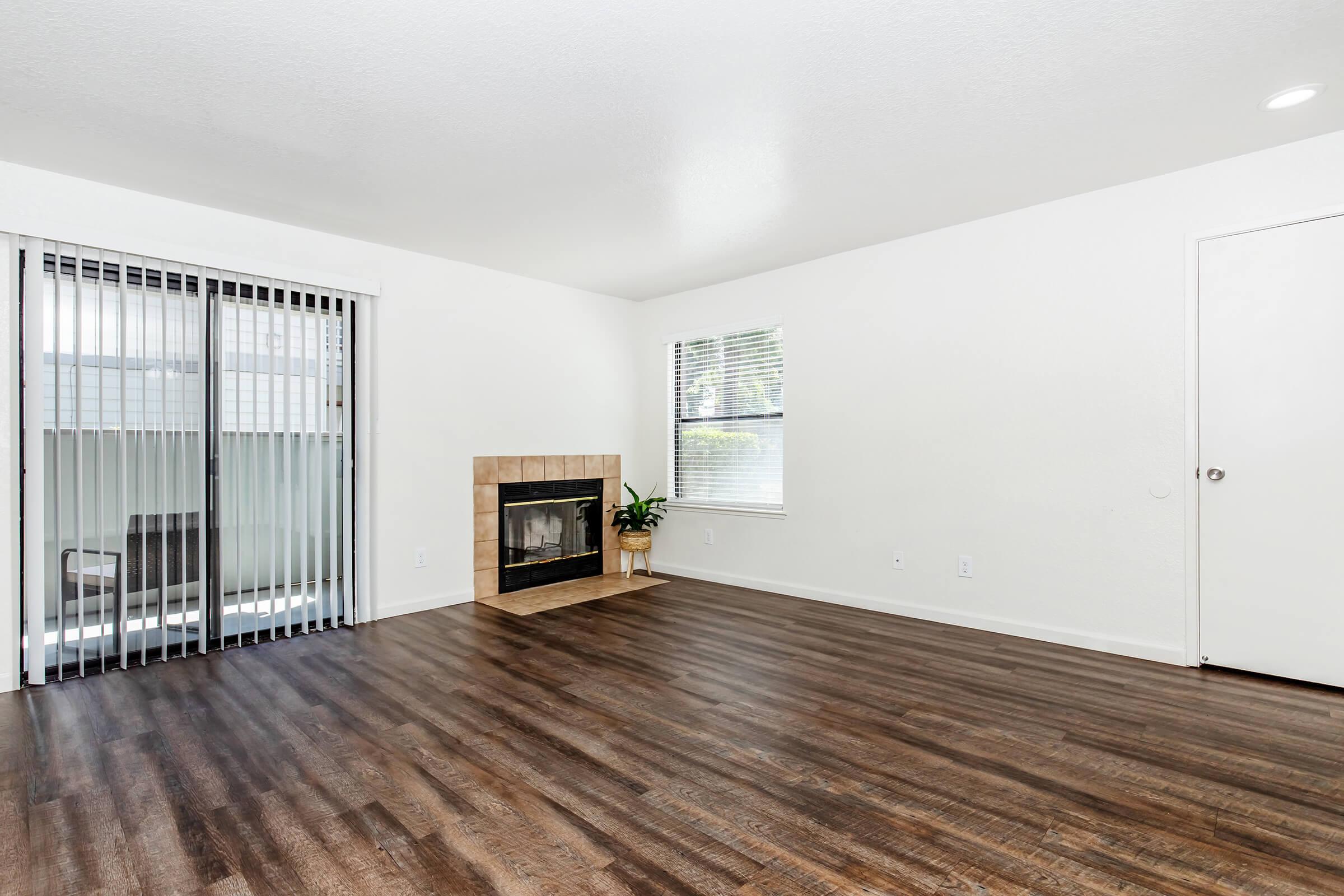 A spacious, empty living room featuring a large window with vertical blinds, a gas fireplace with a tile surround, and a potted plant. The walls are painted white and the flooring is a warm wooden laminate, creating a bright and inviting atmosphere.