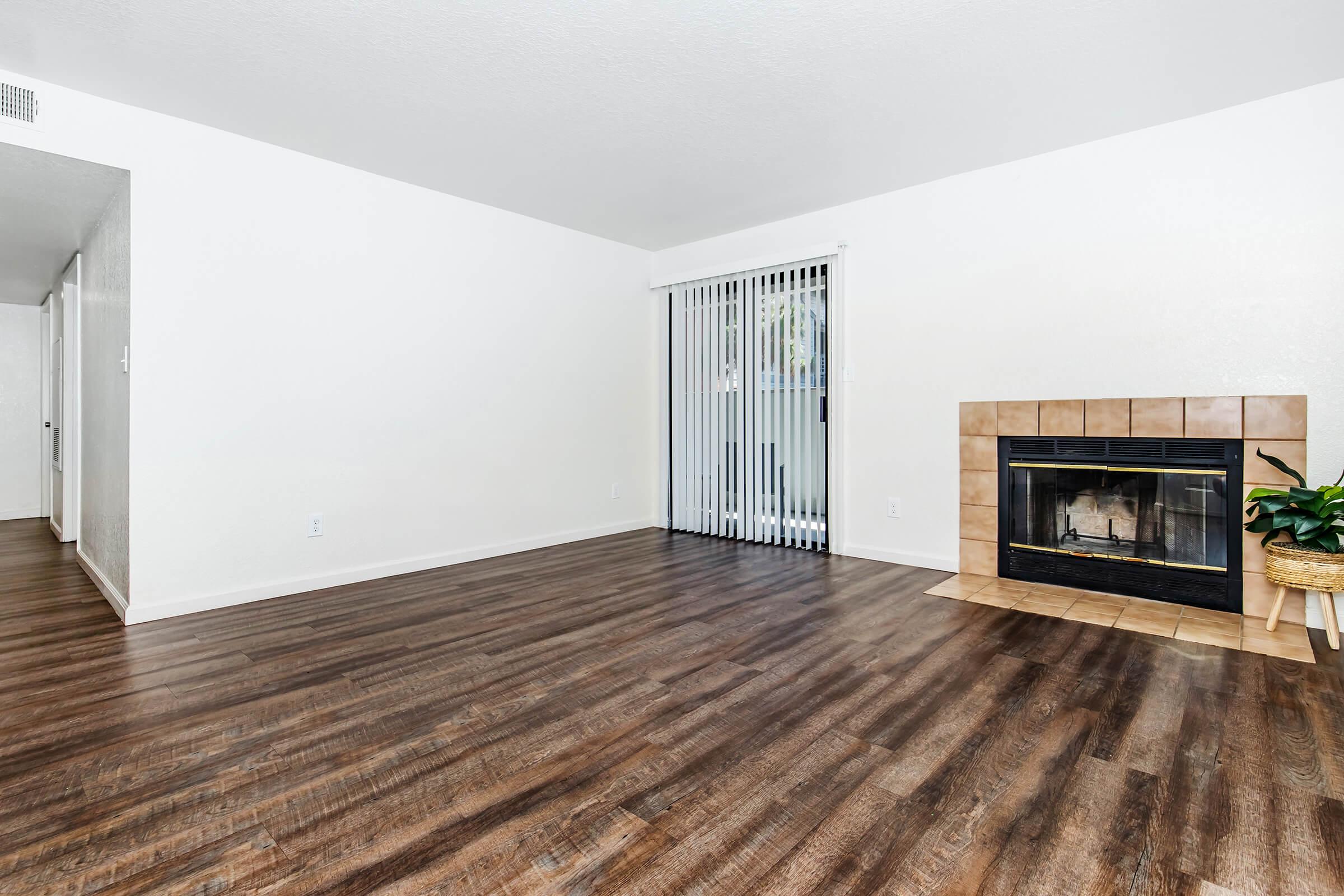 A spacious, empty living room featuring hardwood floors, a tiled fireplace, and large sliding glass doors with vertical blinds. The walls are painted white, and there is a small potted plant near the fireplace. The room is well-lit with natural light.