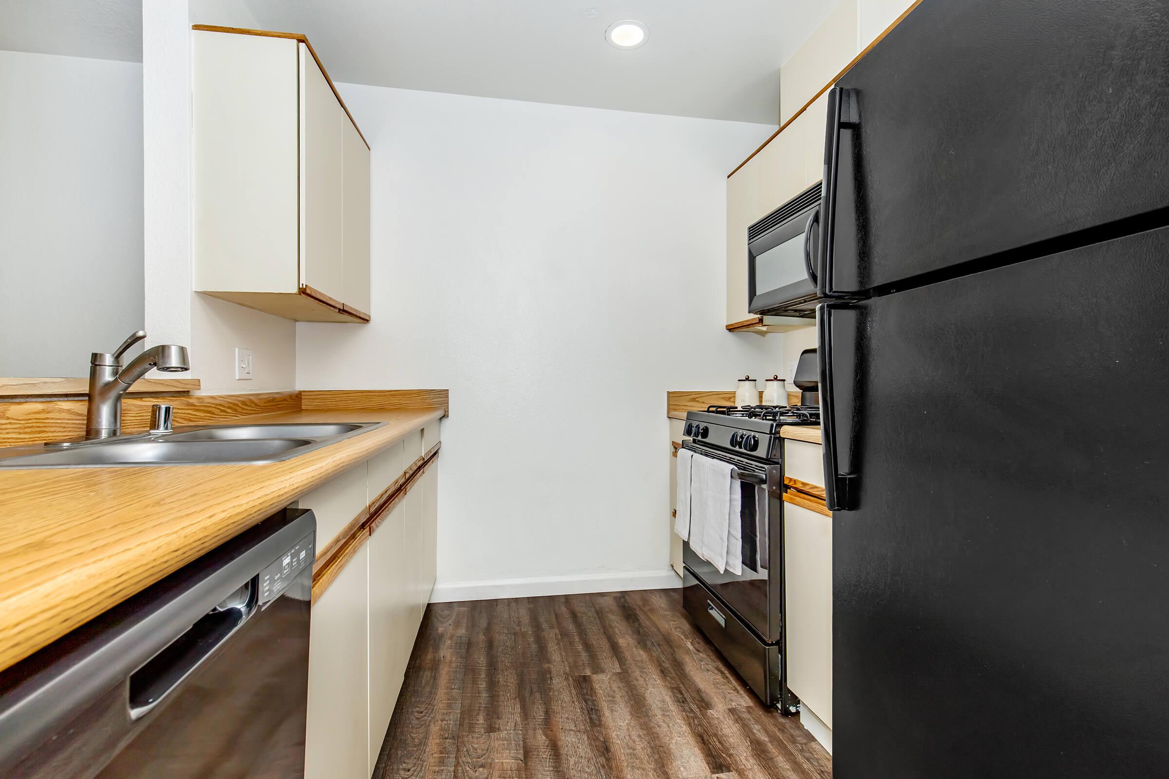 A modern kitchen featuring light-colored cabinetry with wooden countertops, a black refrigerator, and a stainless steel oven. The kitchen has a stainless steel sink and a dishwasher, with a clean and minimalist design. Bright lighting enhances the spacious feel of the room.