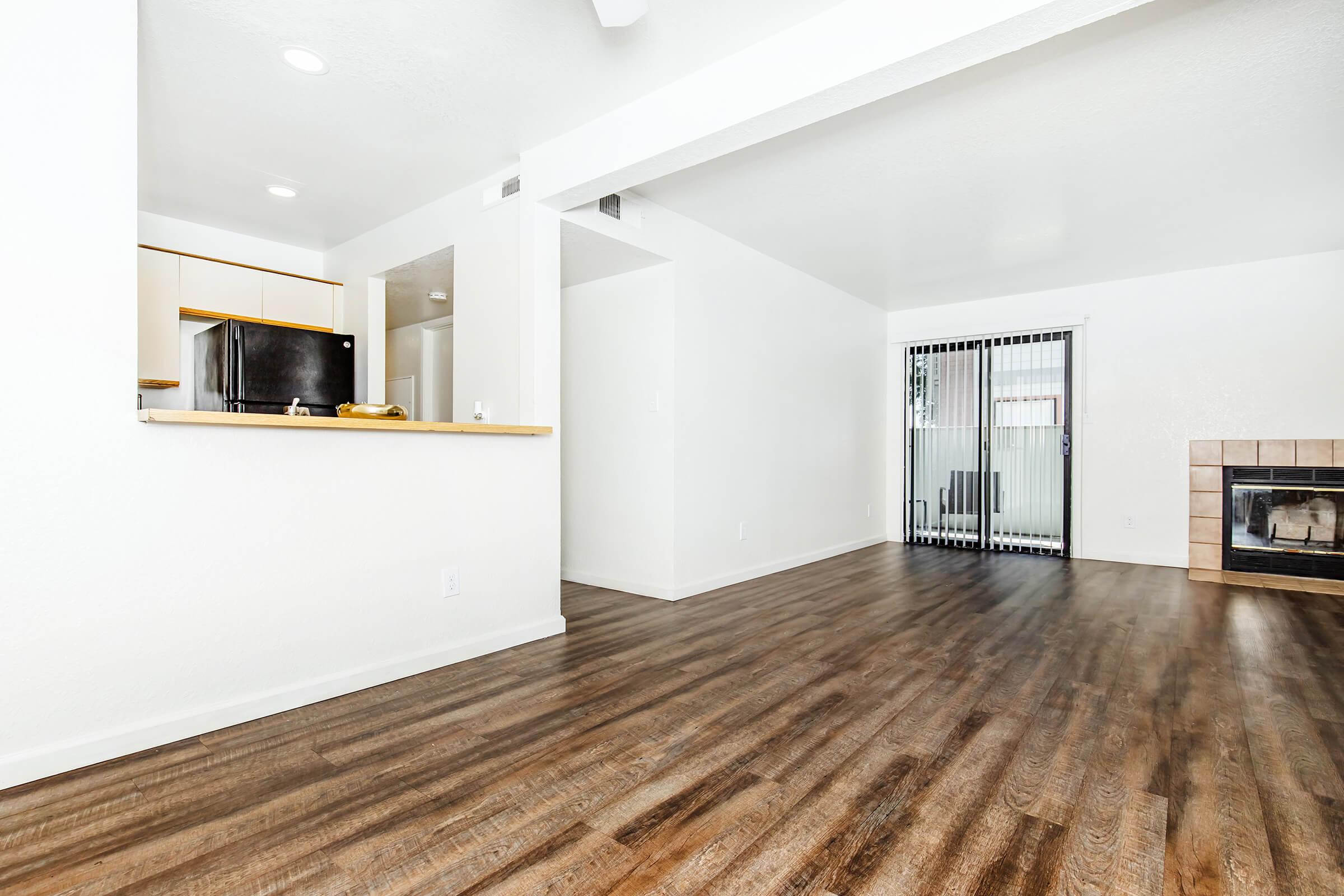 Spacious, modern living room with hardwood floors and a fireplace. To the left, an open kitchen area is partially visible, featuring a black refrigerator and a countertop. The walls are painted white, creating a bright atmosphere. Sliding glass doors lead to an outdoor space, adding natural light to the room.