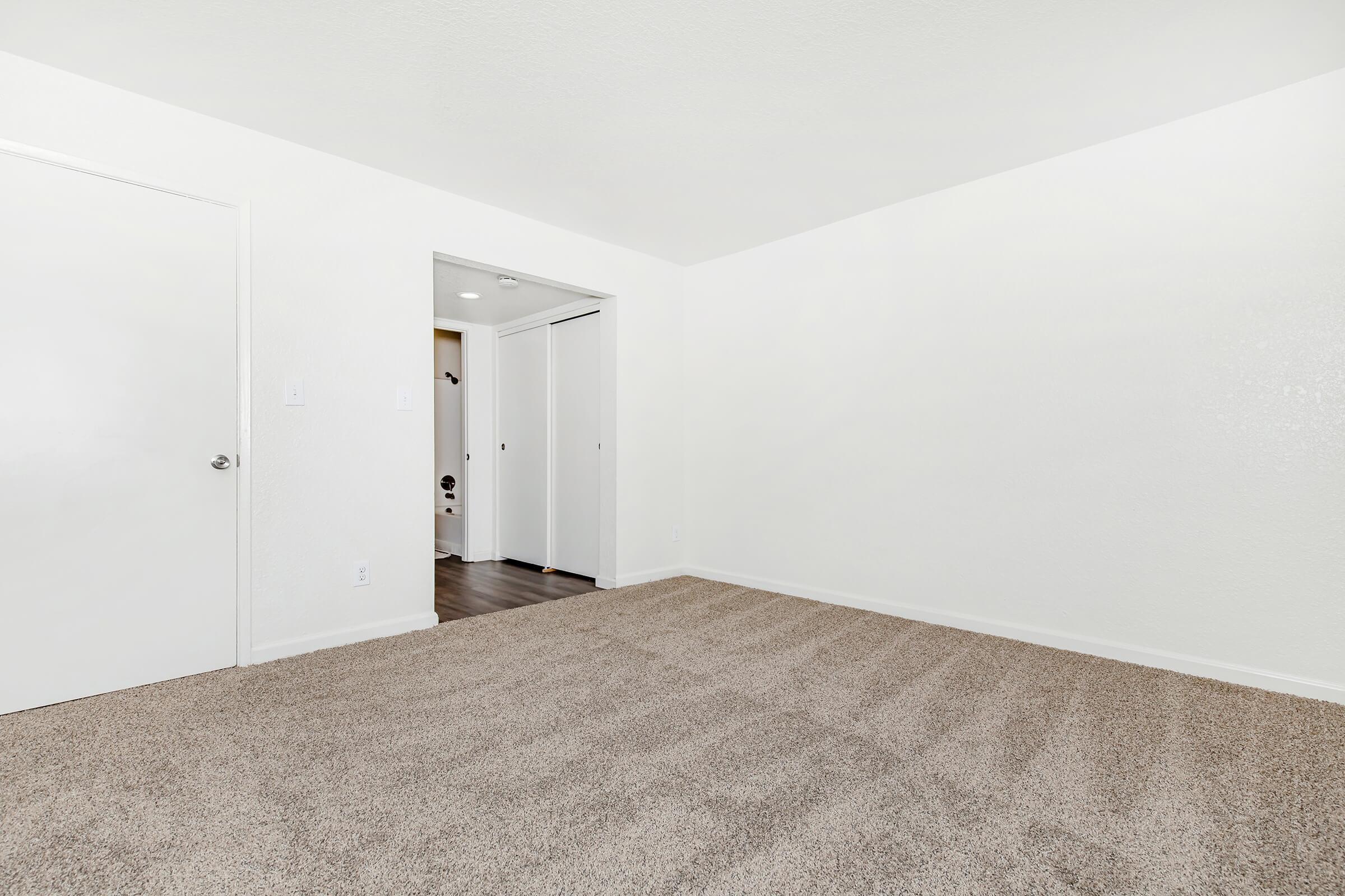 A spacious, empty room with light-colored walls and a plush, beige carpet. There are two doors on the left side leading to adjacent spaces, and the floor is clean and well-maintained. The overall atmosphere is bright and inviting, perfect for personalizing or decorating.