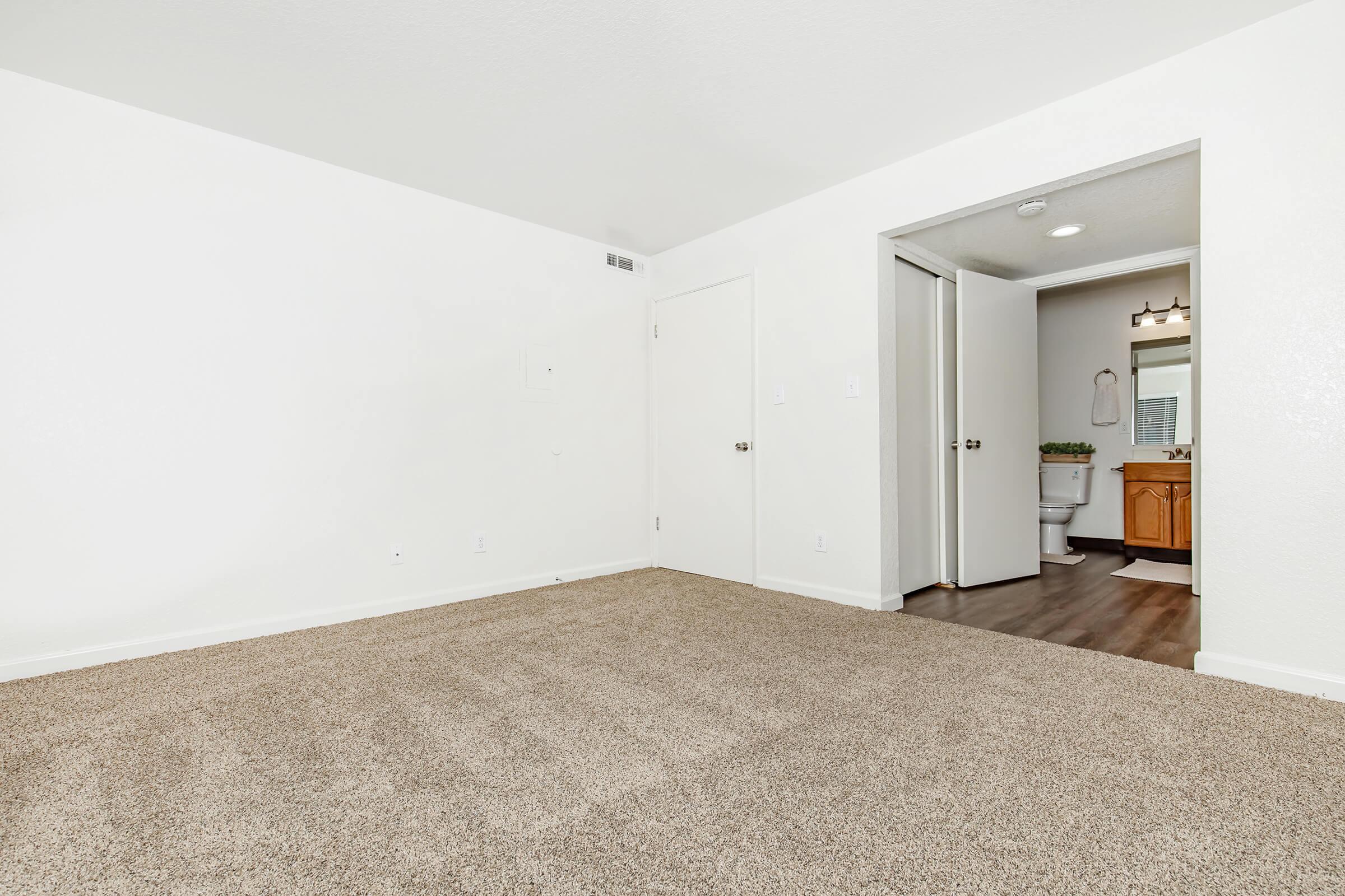 A spacious, empty living room with light beige carpet and white walls. On the right, a doorway leads to a bathroom featuring wooden cabinets, a mirror, and white fixtures. Natural light illuminates the area, creating a bright and open atmosphere.