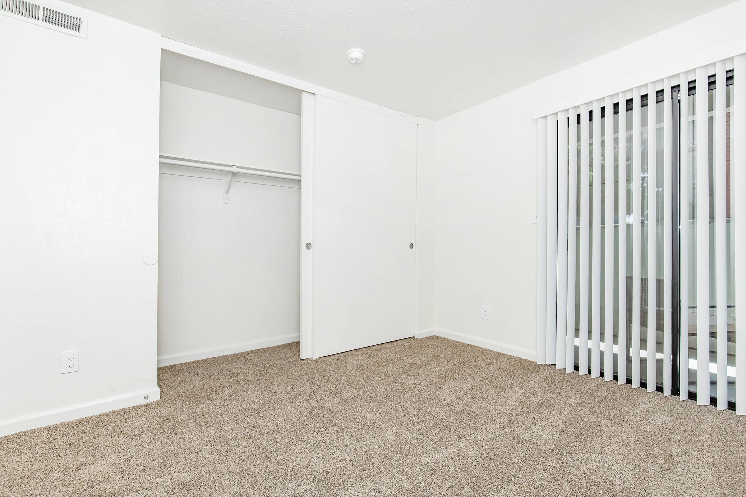An empty room with light beige carpet, white walls, and a sliding glass door featuring vertical blinds. There is a closet with sliding doors on the left side of the image, and the overall ambiance is bright and minimalistic.