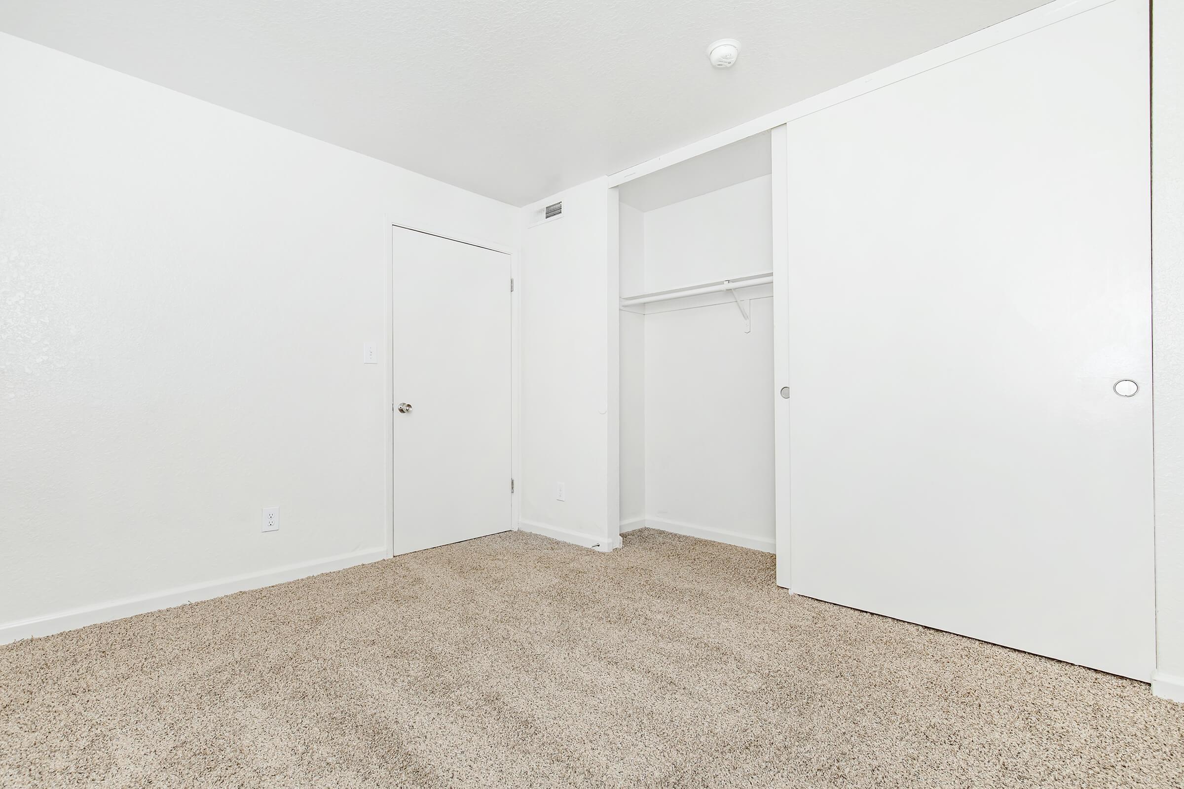 A clean, empty room with light-colored walls and carpet. There is a closet with sliding white doors and a single door leading out of the room. The space is well-lit, featuring minimal décor, emphasizing a fresh and spacious atmosphere.